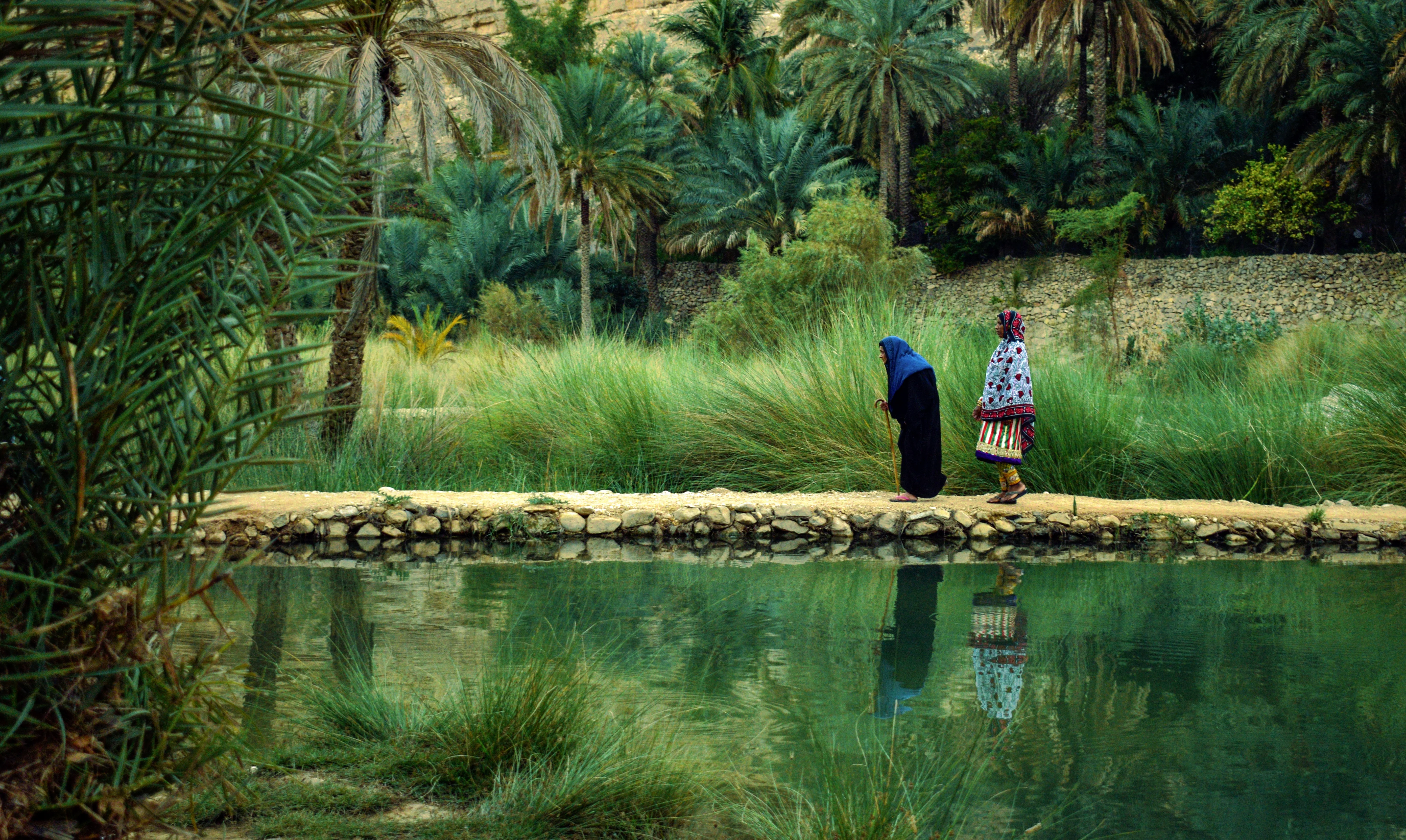 Two women walking beside falaj waterway in lush Wadi on an Oman tour.