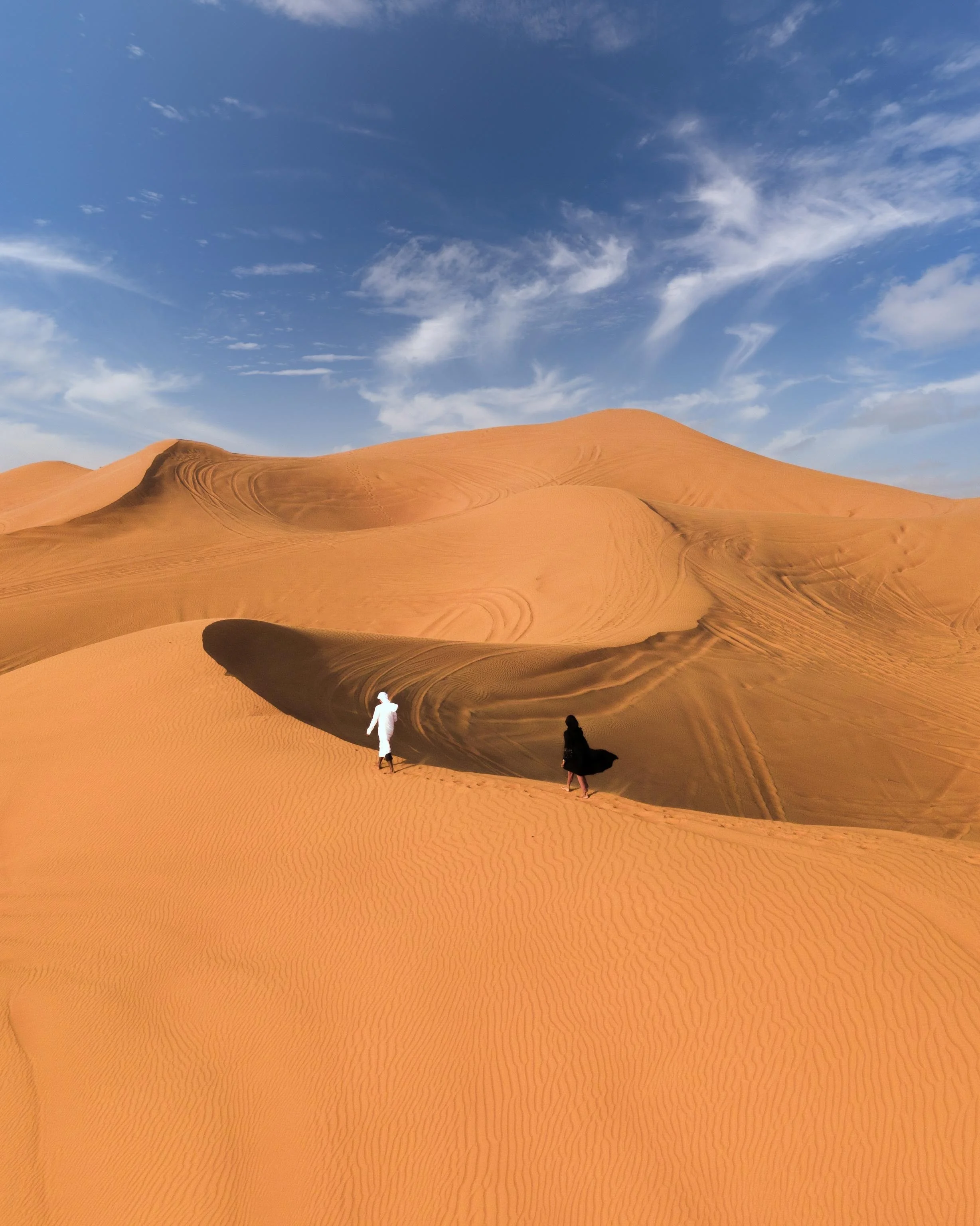 Two people walking across tall desert dunes under blue sky