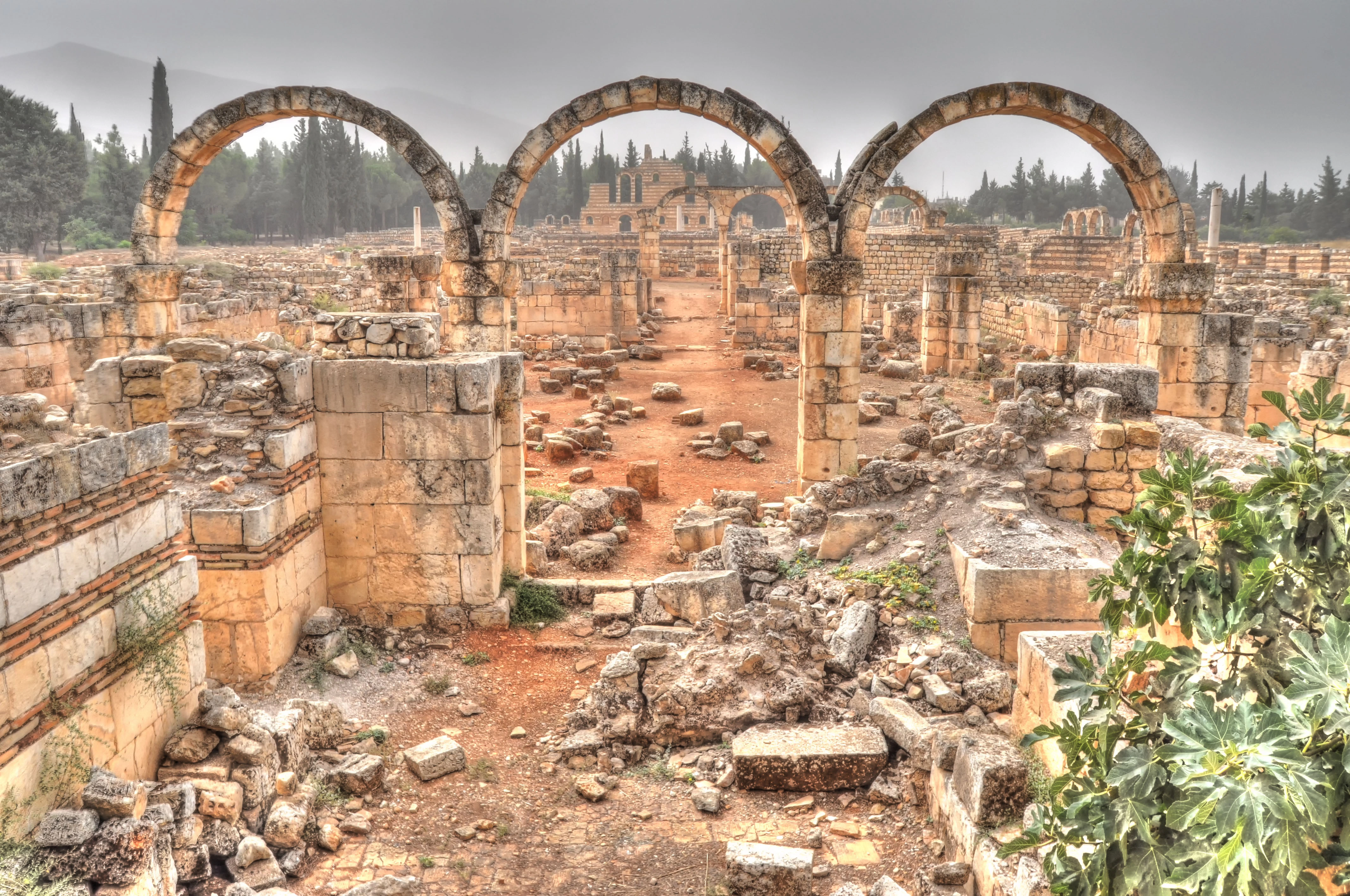 Three stone arches and ancient ruins of Anjar with foggy mountains in the background.