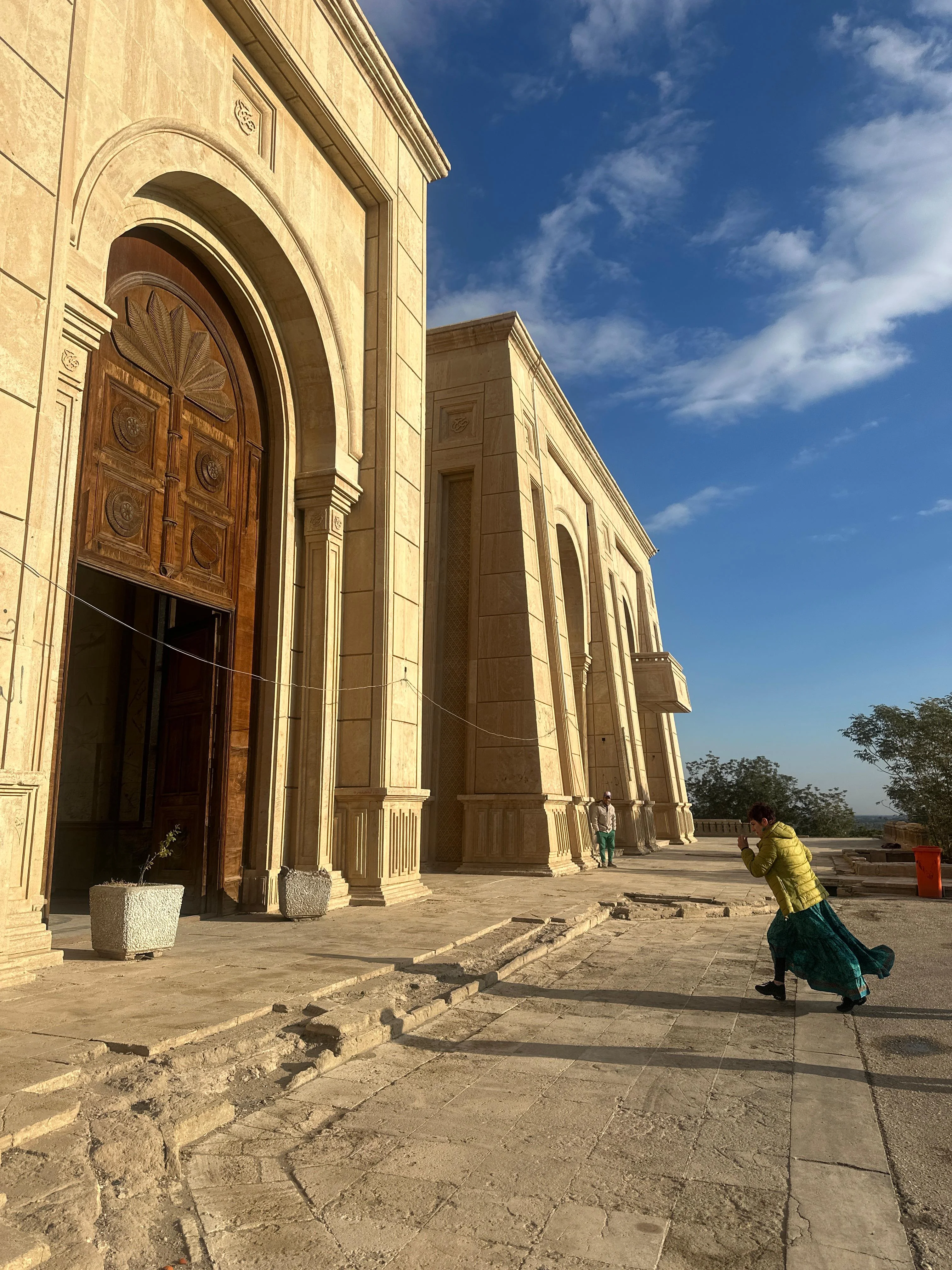 Woman walking outside a grand sandstone building in Iraq with arched wooden doors and blue sky