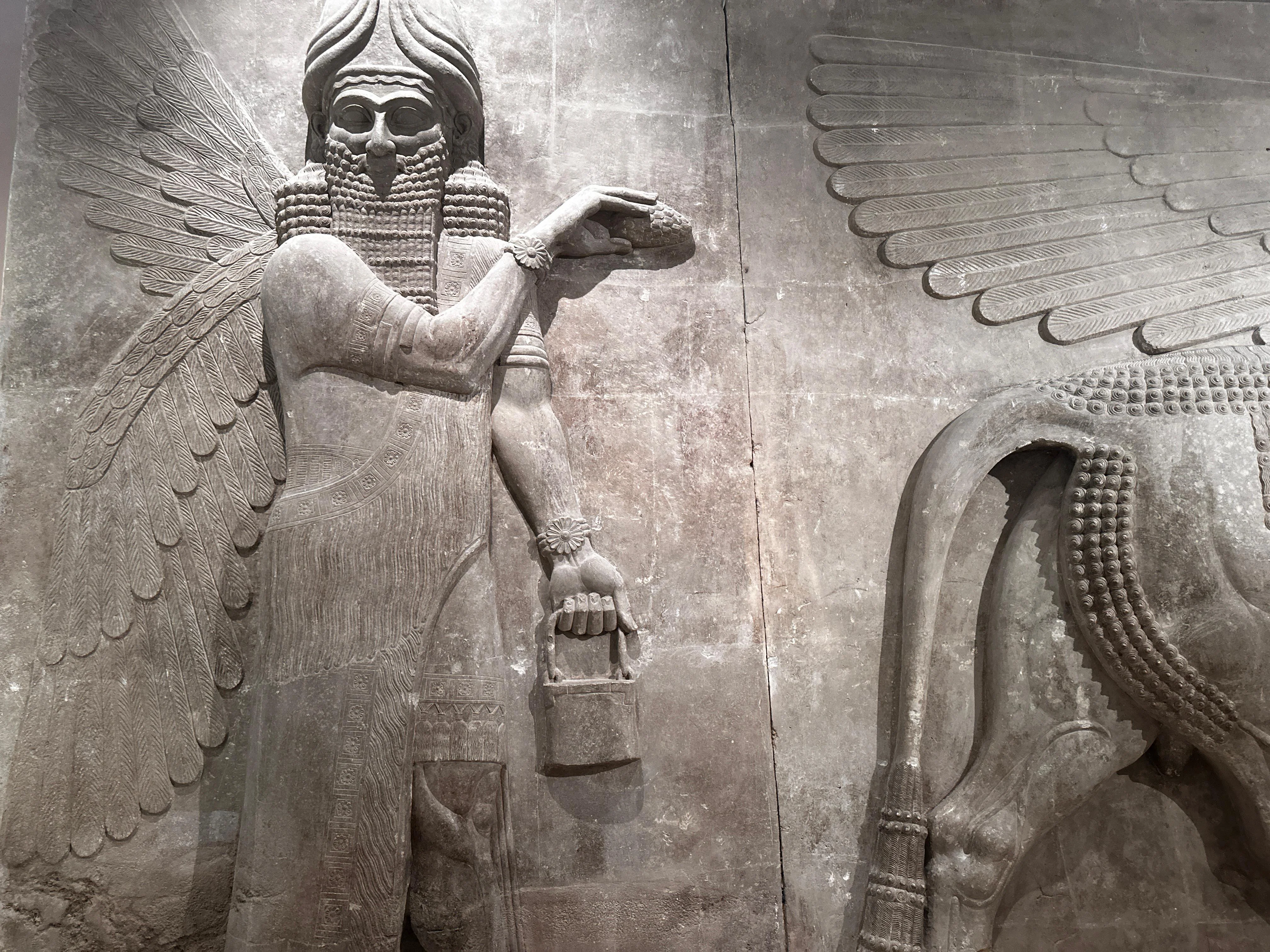 Winged Assyrian deity carved in stone holding a bucket and pinecone, symbolizing protection