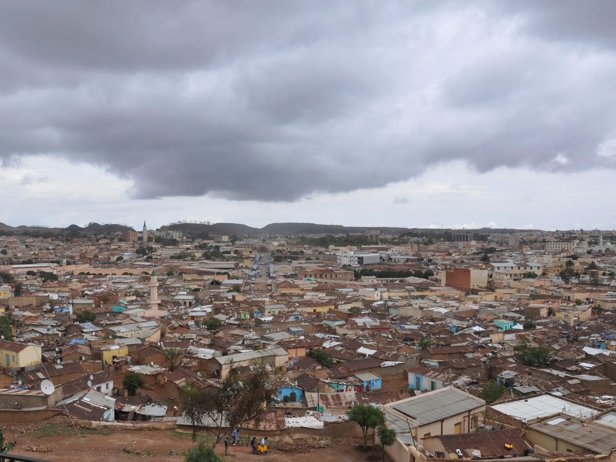 Panoramic view over Eritrean city with dense low-rise homes, mosque minarets and mountains beneath storm clouds