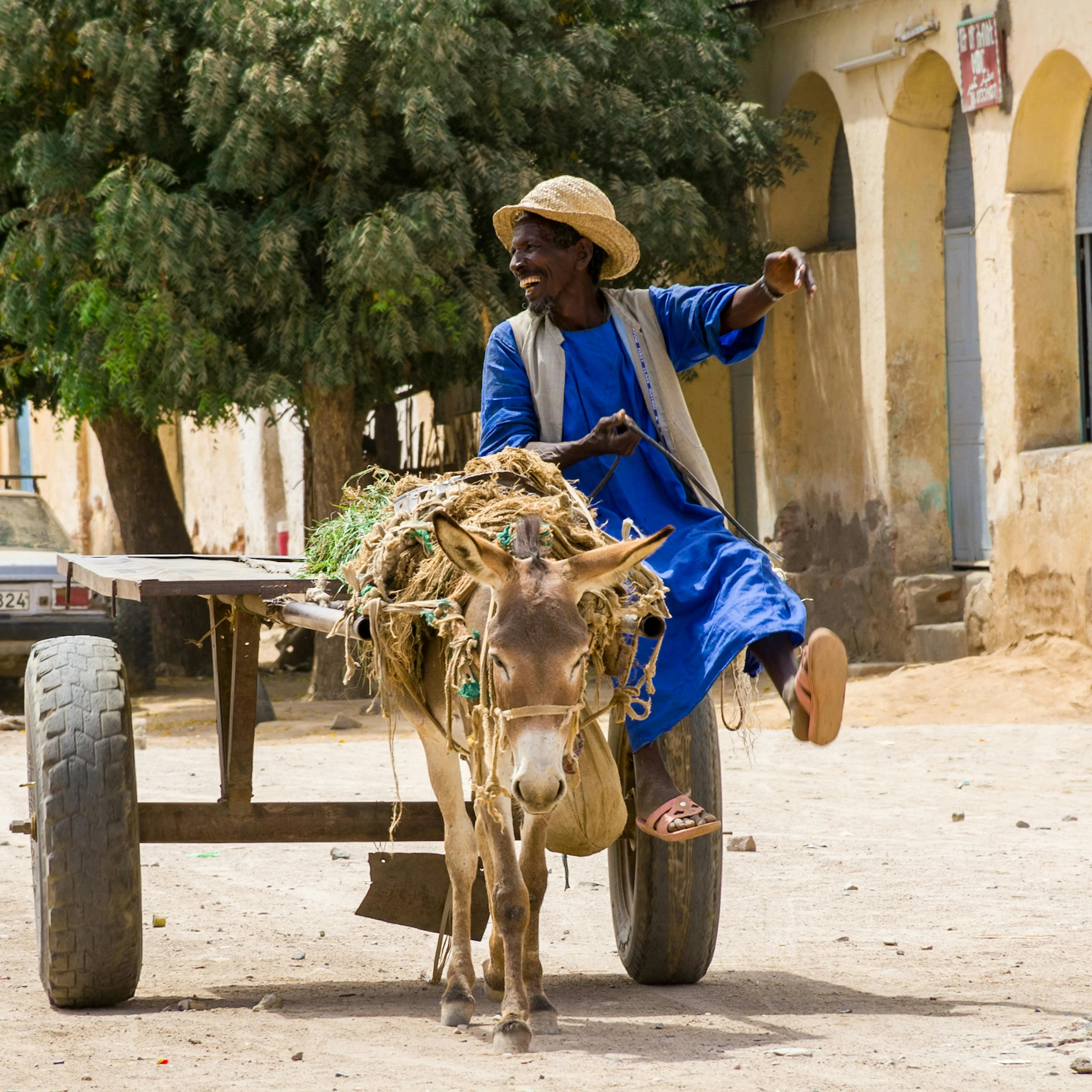Eritrean man in blue tunic and straw hat rides a donkey cart laden with hay through a dusty street in an old town
