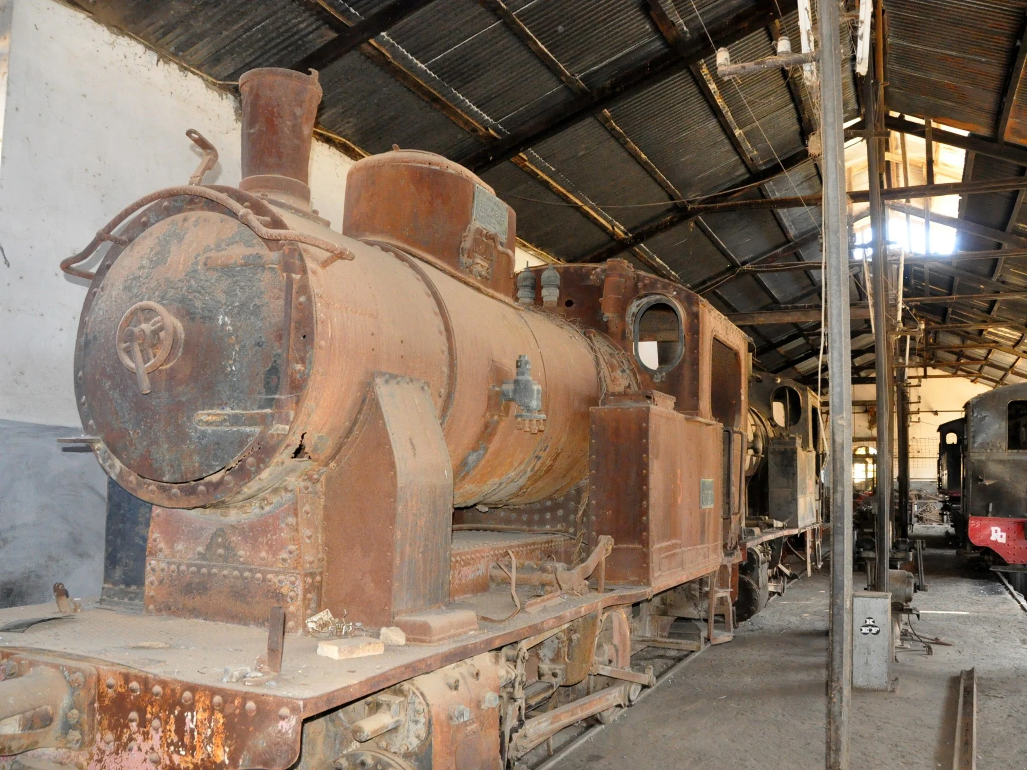 Rusted steam locomotive in Eritrean railway workshop, with other vintage trains under a corrugated metal roof