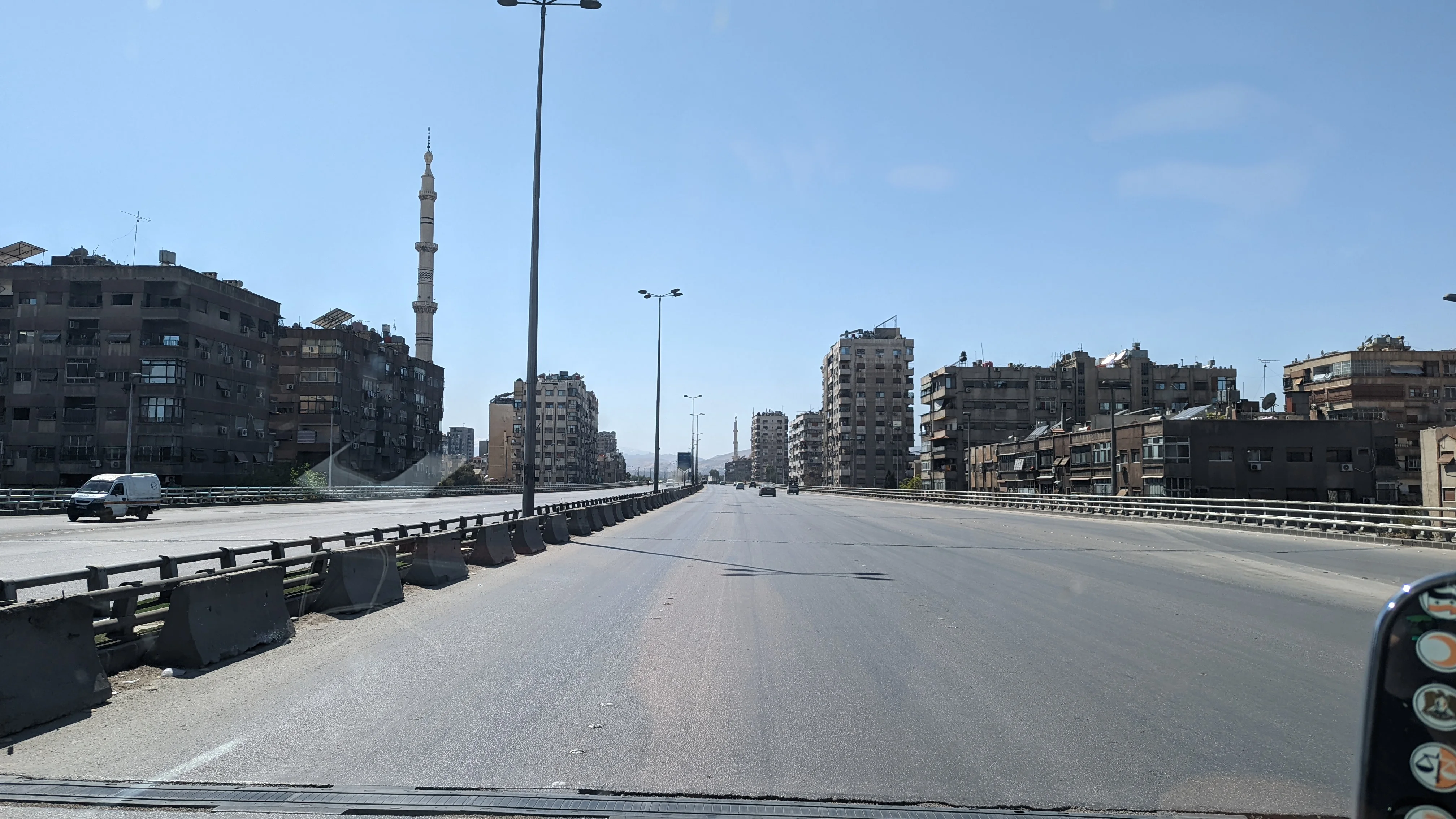 Wide highway in Damascus lined with mid-rise buildings and a tall mosque minaret in the distance