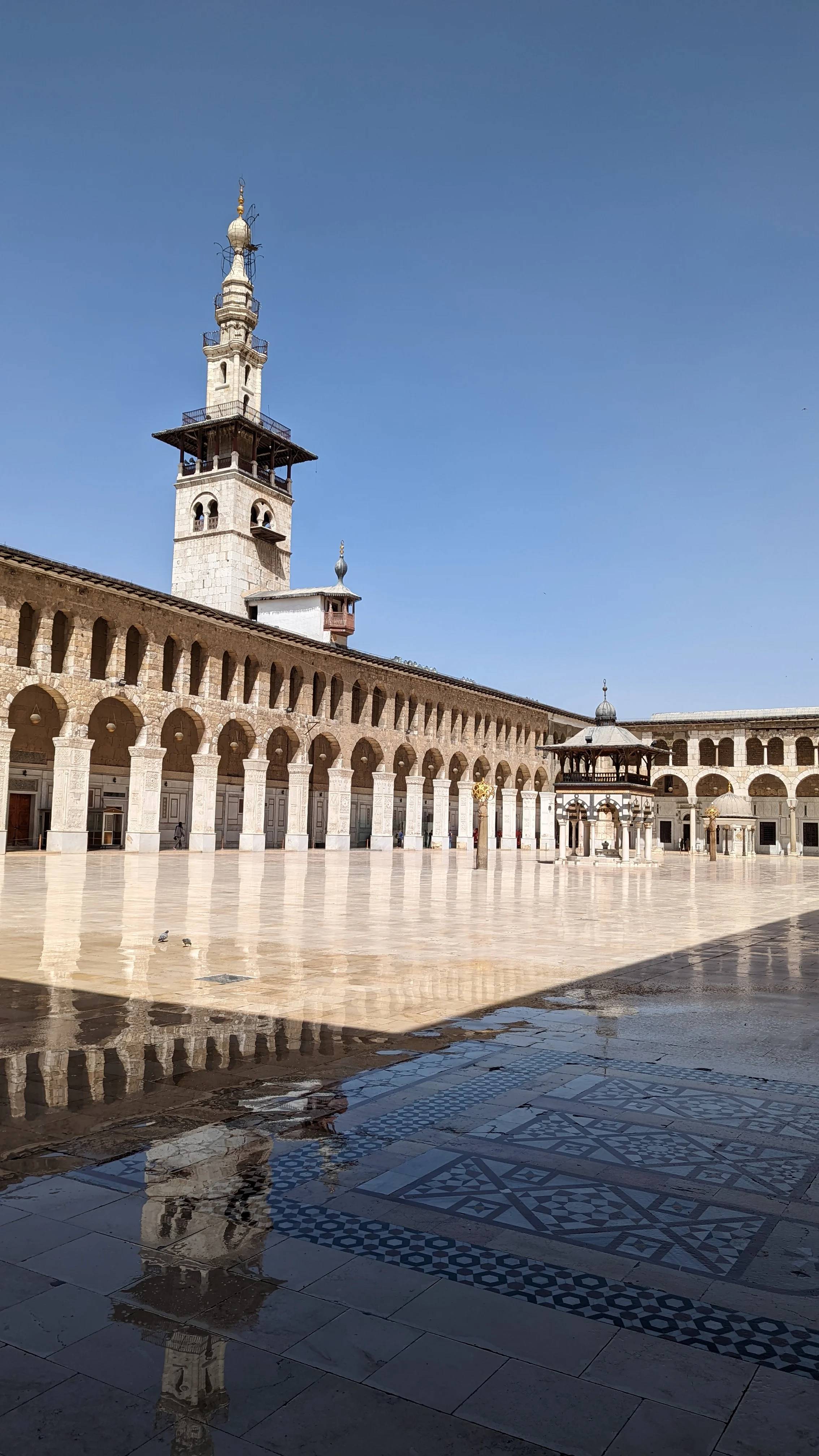 Courtyard of the Umayyad Mosque with minaret and arched colonnades reflected in a wet tiled floor