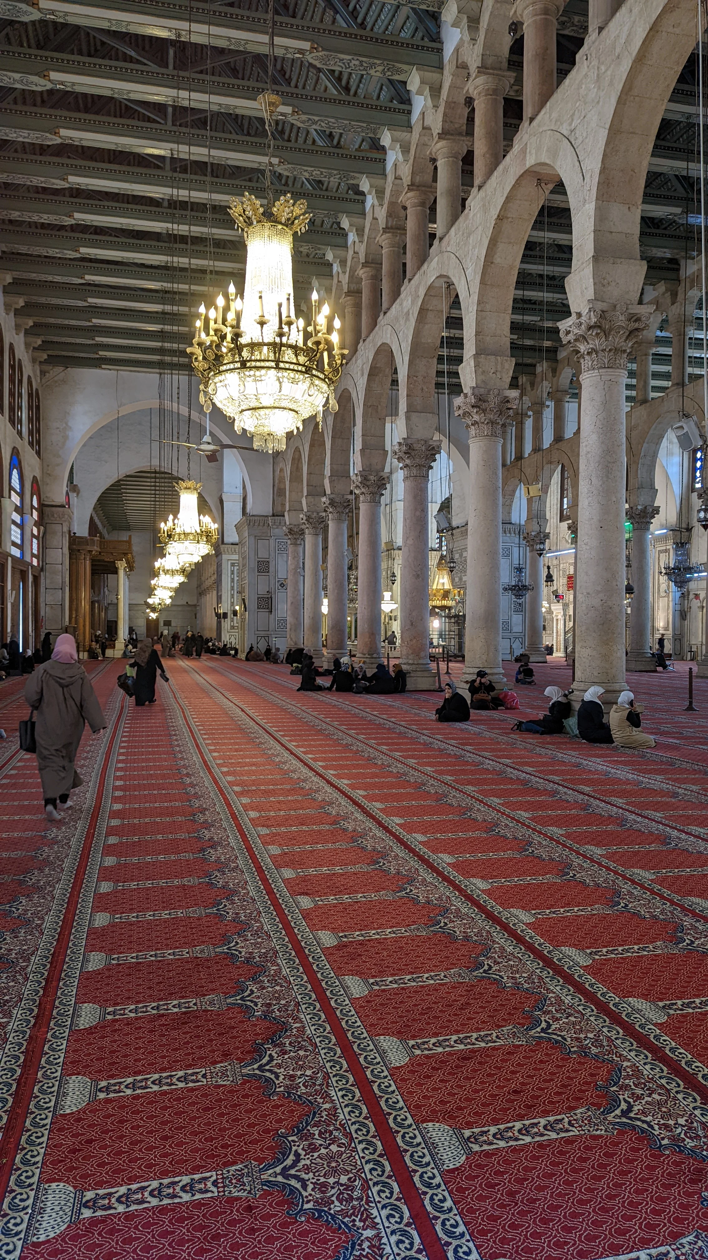 Interior of the Umayyad Mosque with red prayer carpet, chandeliers, and people seated in prayer