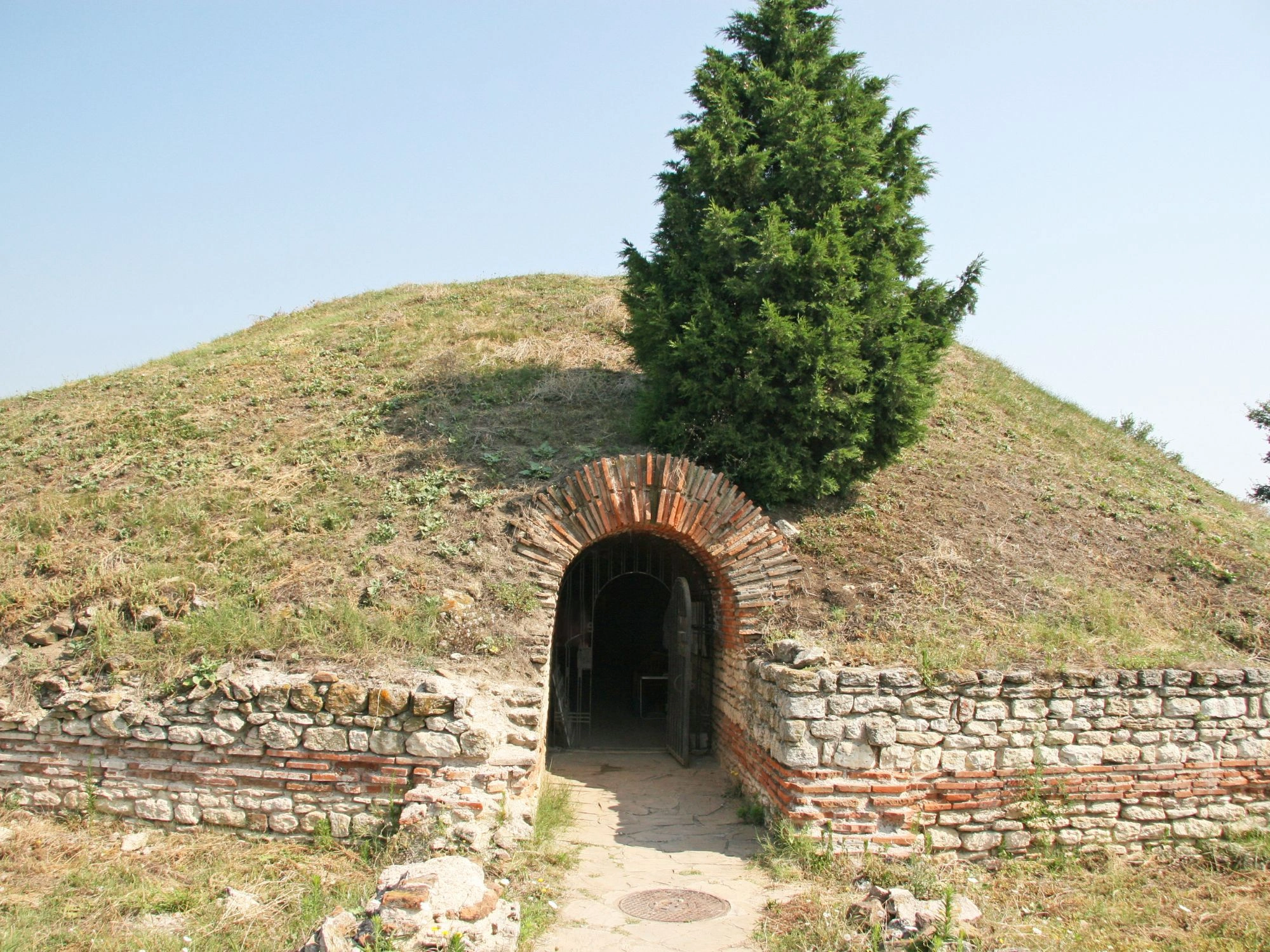 Grass-covered Thracian burial mound with stone entrance and brick arch.