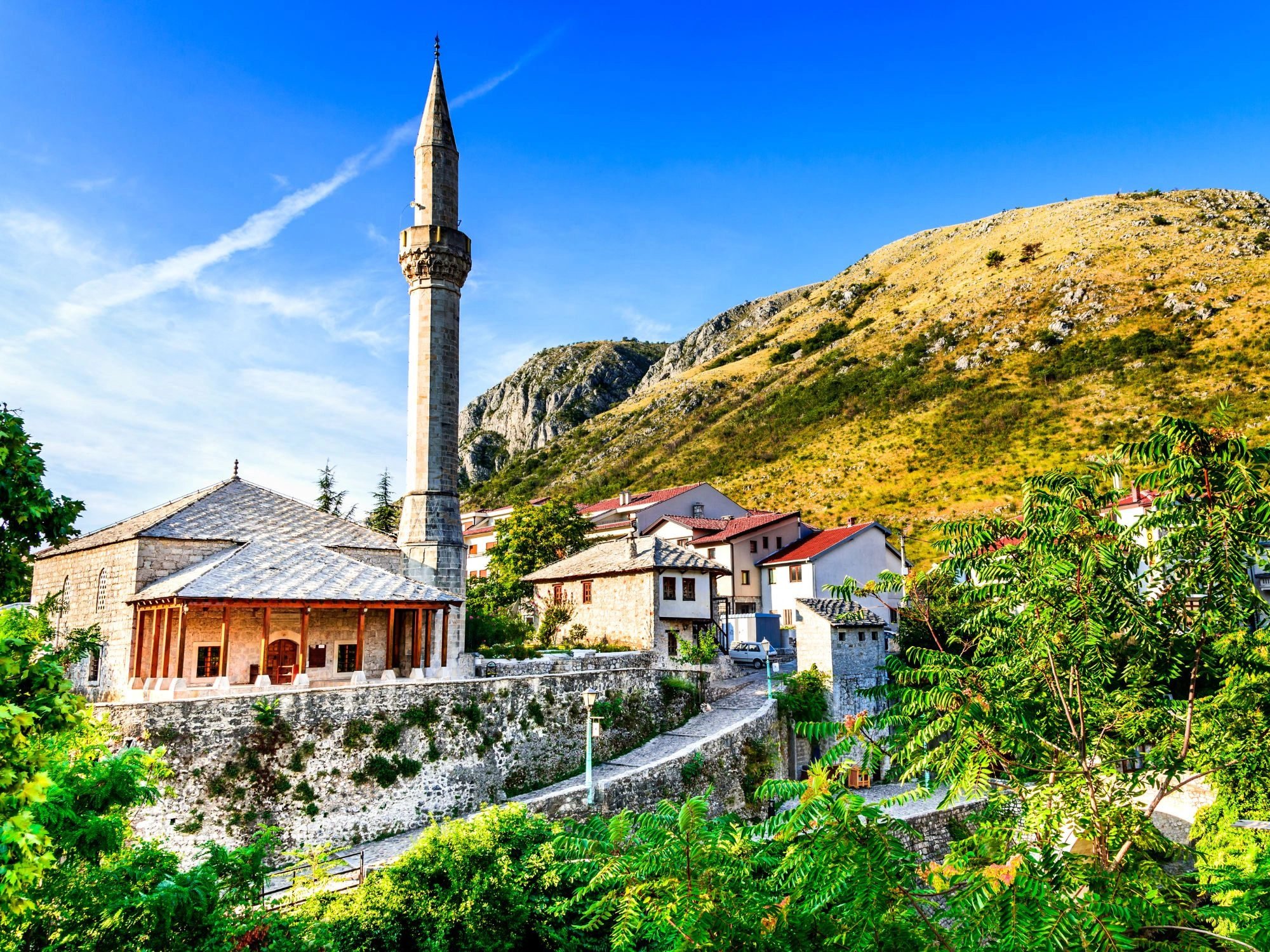 Traditional stone mosque with a tall minaret and rustic houses in Mostar, Bosnia and Herzegovina.
