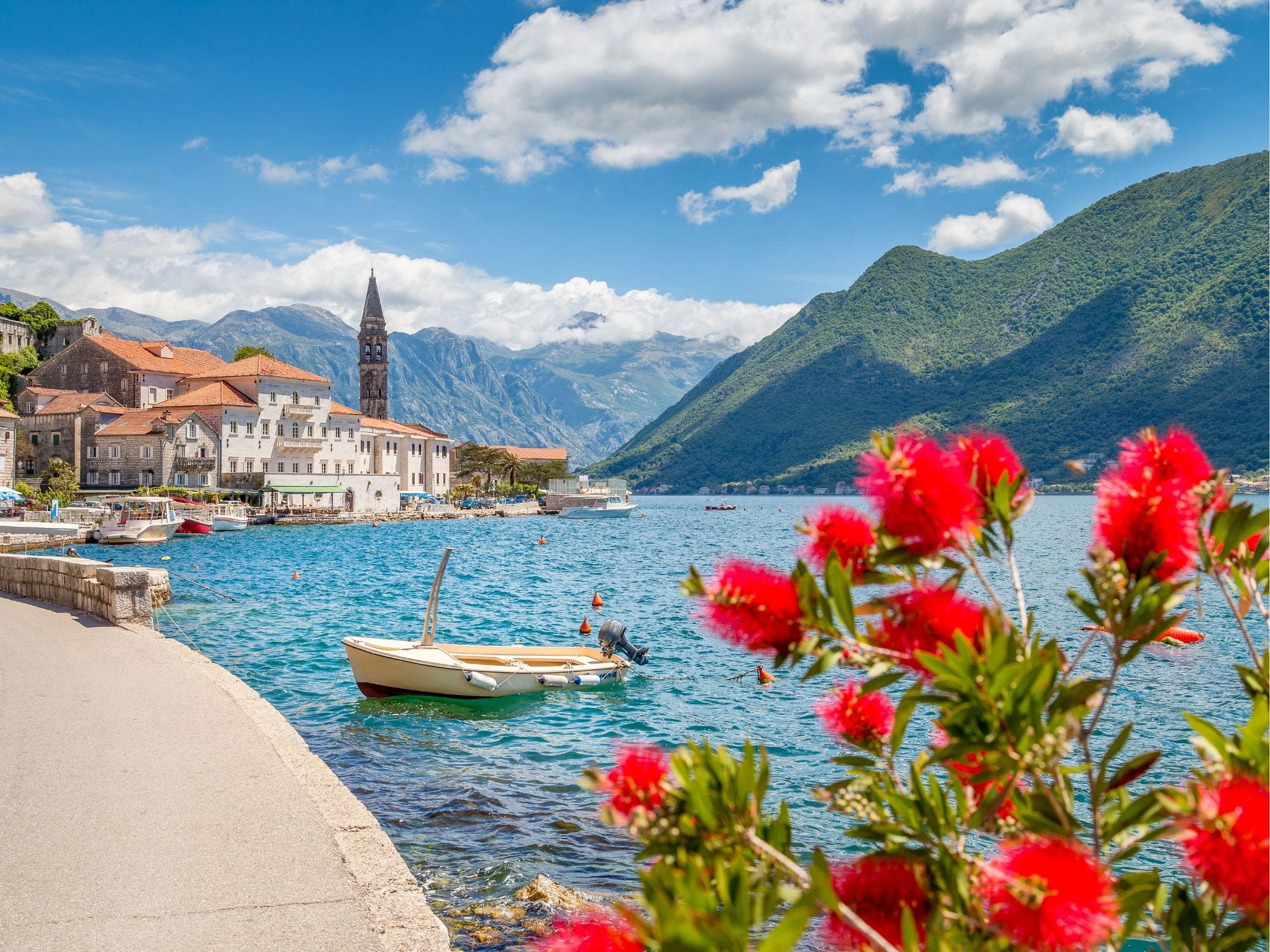 Scenic waterfront of Perast, Montenegro, with red flowers, stone houses, and lush mountains.