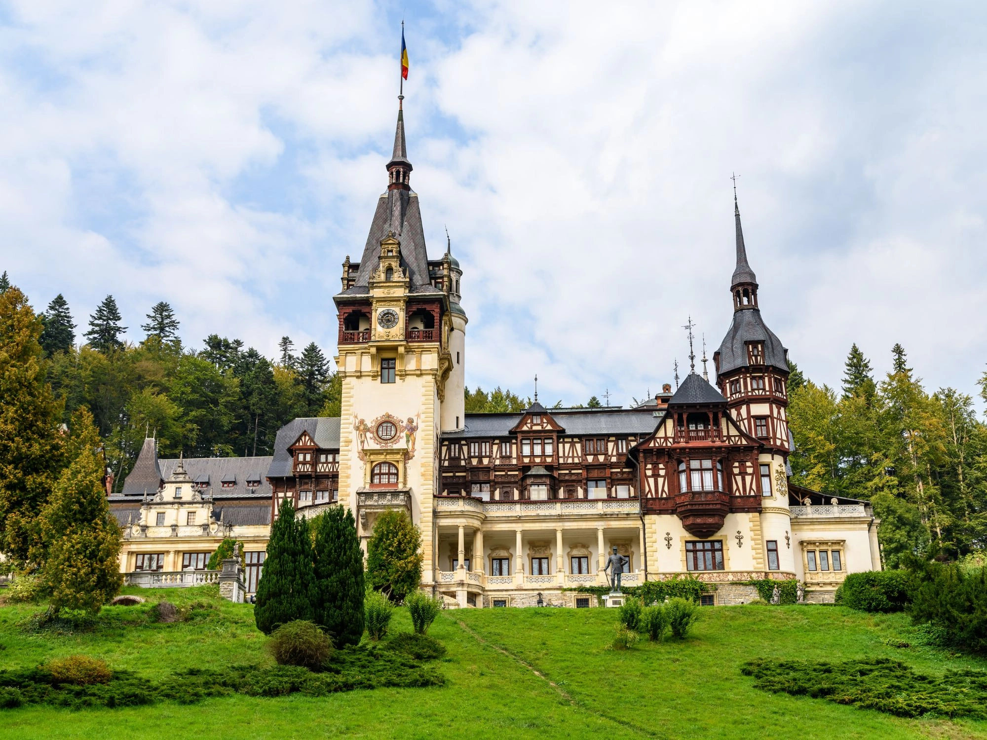 Ornate Romania Peles Castle in Sinaia with spires, gardens and Romanian flag on top.