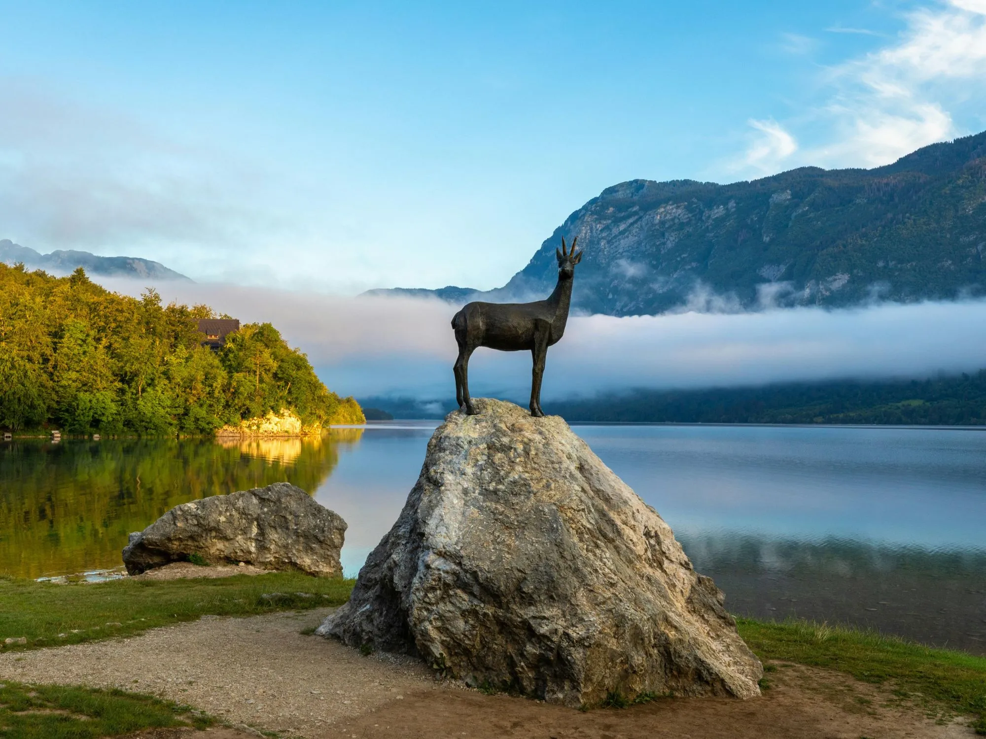A scenic view of Lake Bohinj in Slovenia with the iconic Zlatorog chamois statue standing on a rock at the water's edge, mist hovering over the lake, and forested mountains in the background.
