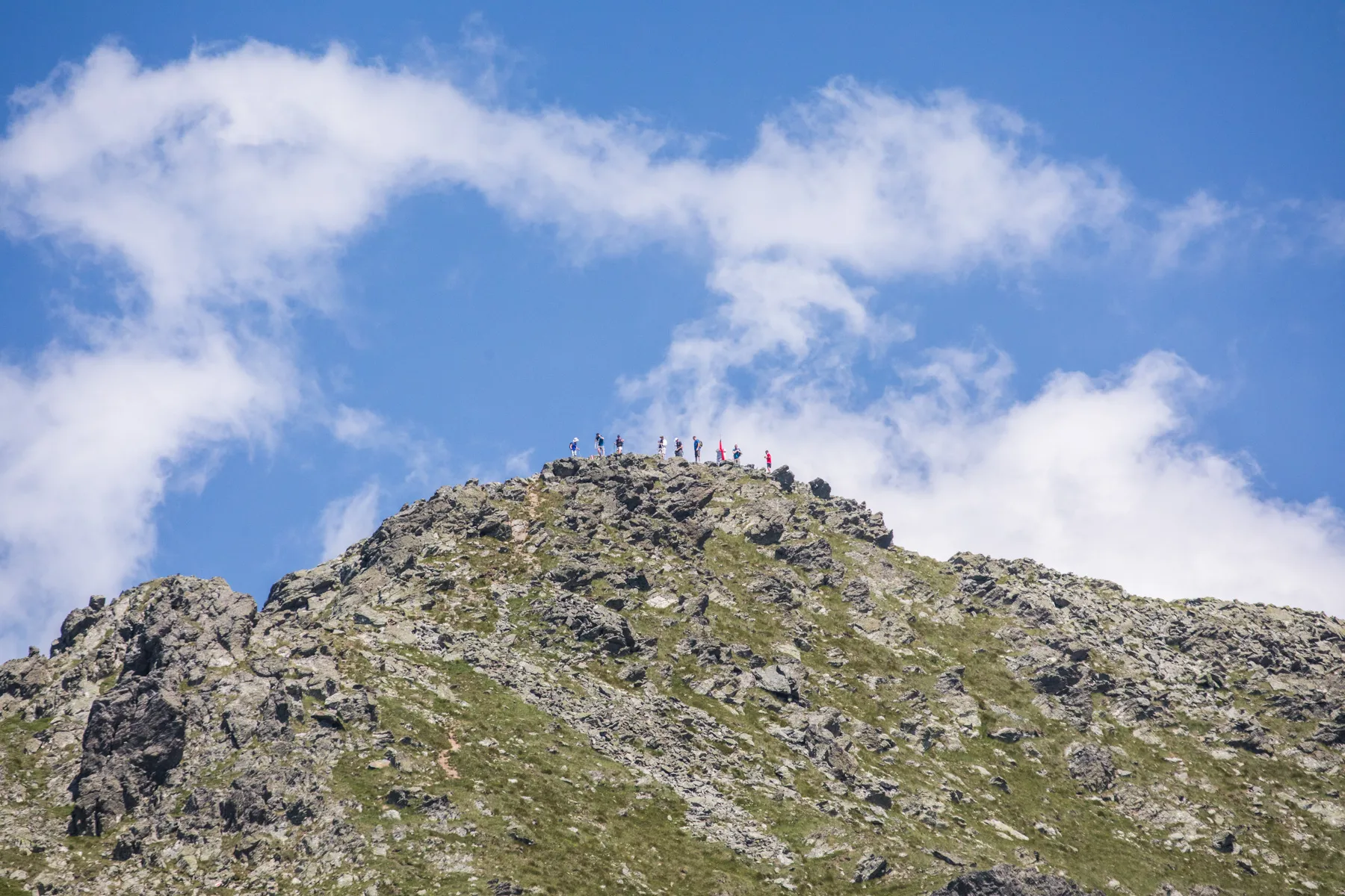 Distant hikers reach the rocky summit of Gjeravica Peak under bright skies along the Via Dinarica in Kosovo