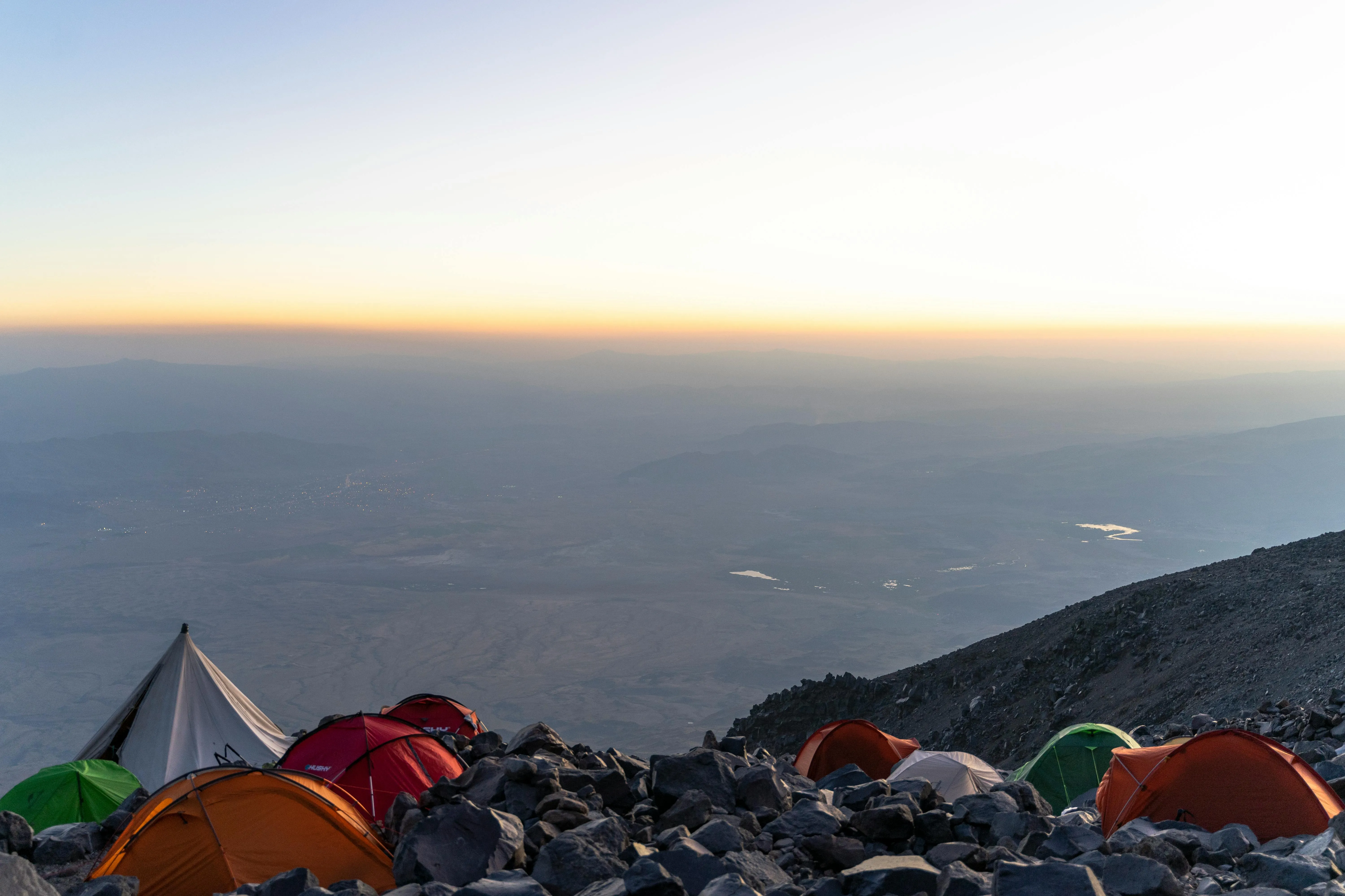 A group of tents pitched on a rocky slope, overlooking a vast landscape at dusk.