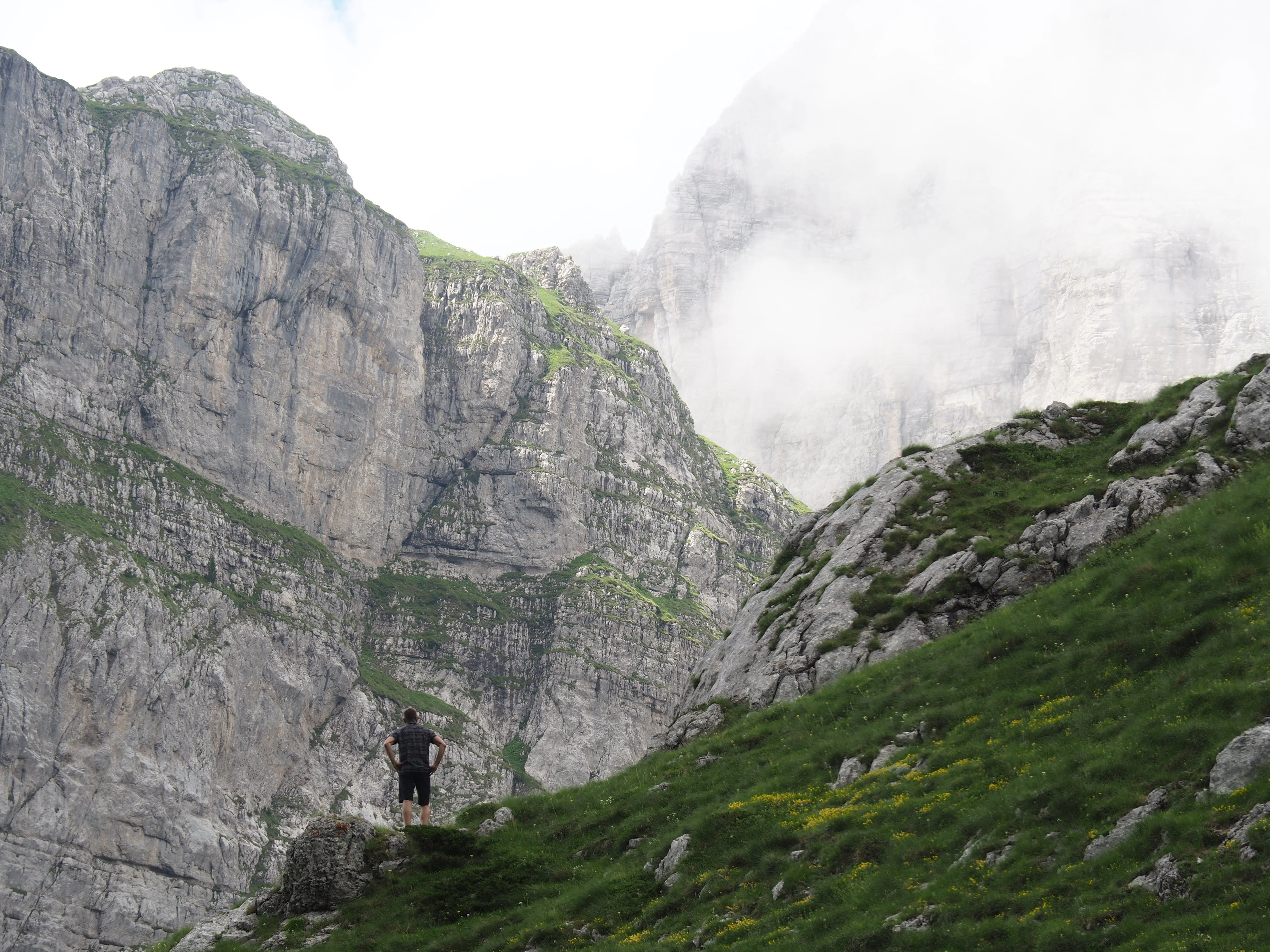 A lone hiker stands atop a grassy ridge, gazing at the towering, mist-covered cliffs of the Albanian Alps.