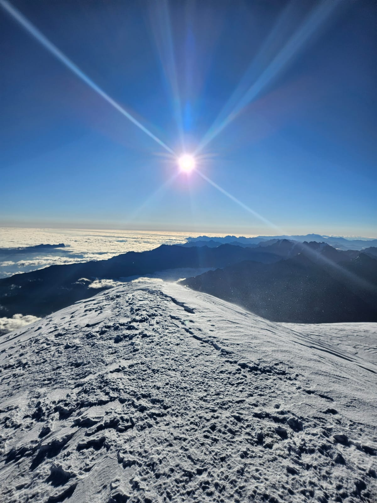 Snow-covered mountain summit rising above a sea of clouds under bright sunburst in clear blue sky