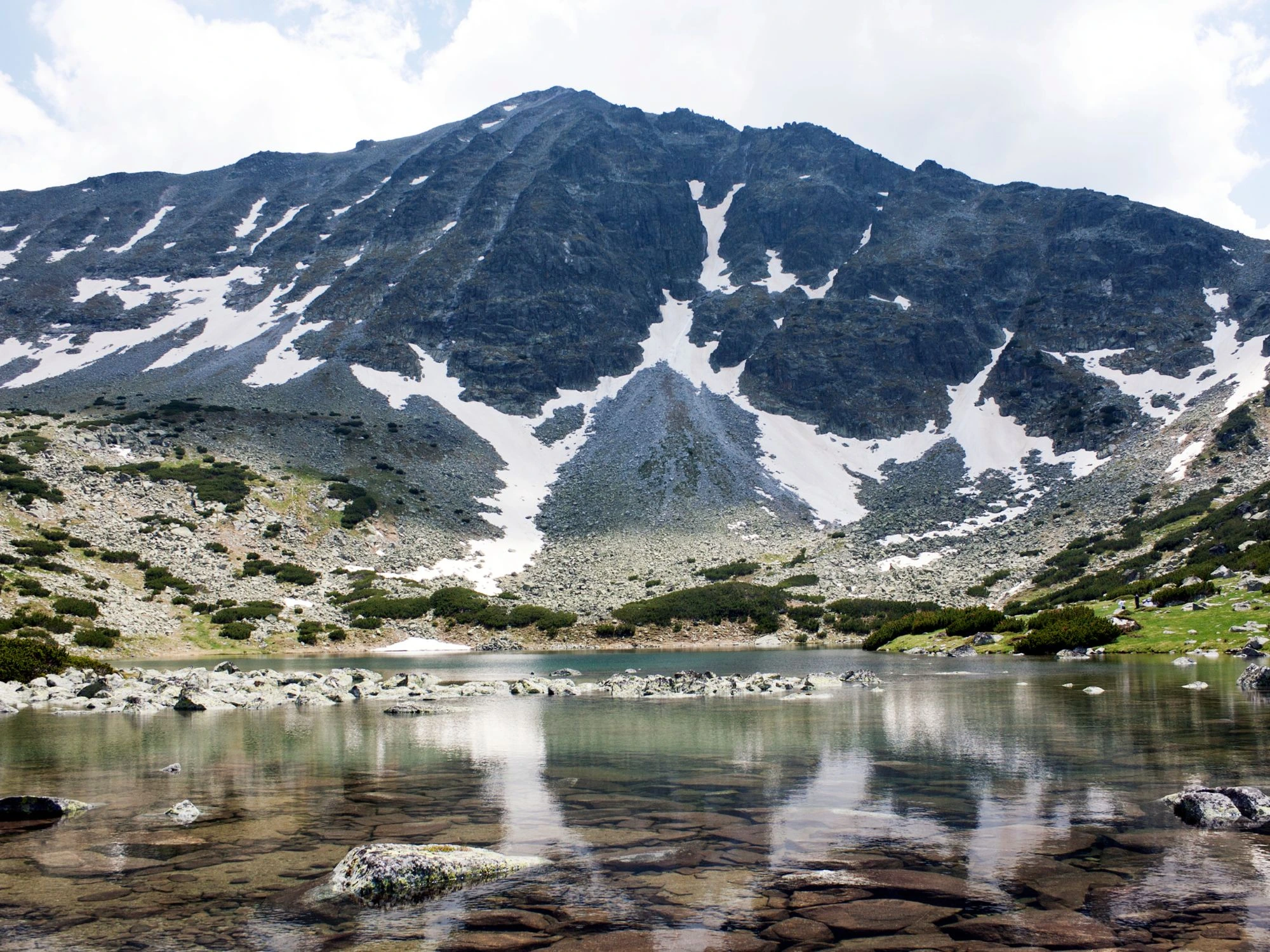 A serene mountain lake in Rila, Bulgaria, reflecting the rugged, snow-streaked slopes of the surrounding peaks.