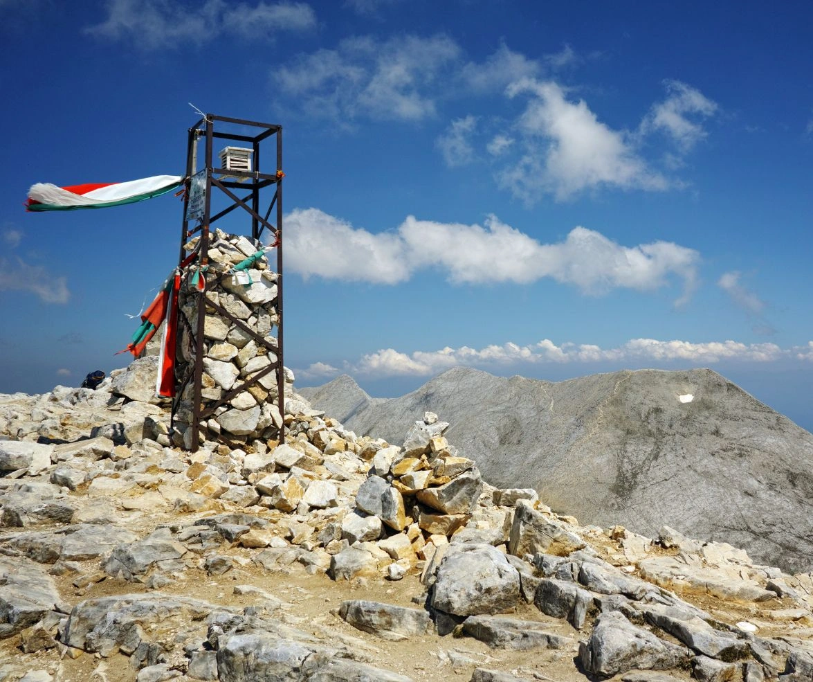 Trig point cairn draped with Bulgarian flag on Mount Vihren summit overlooking rocky Pirin peaks