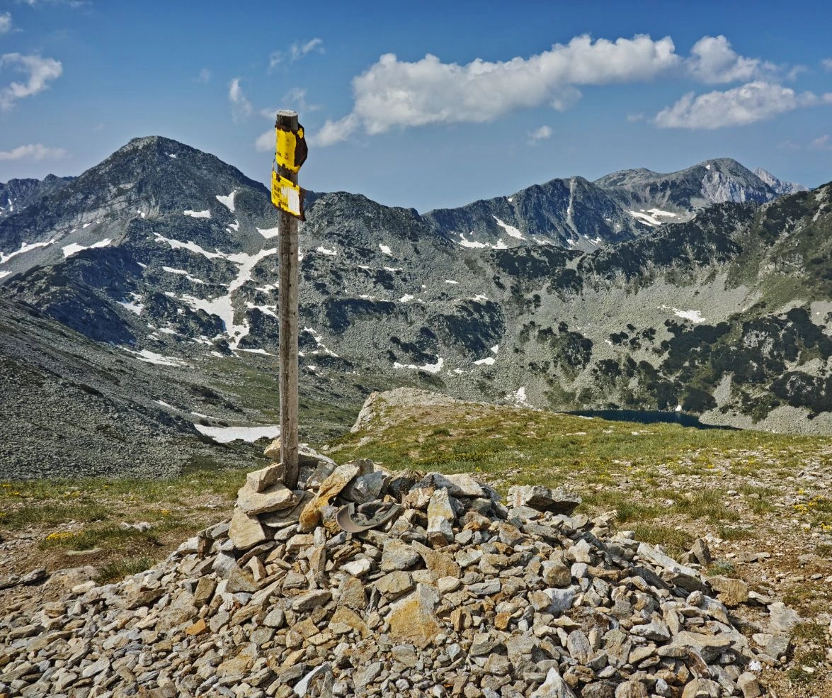 Rock cairn with yellow trail marker on Vihren hiking route overlooking rocky Pirin peaks and alpine lakes