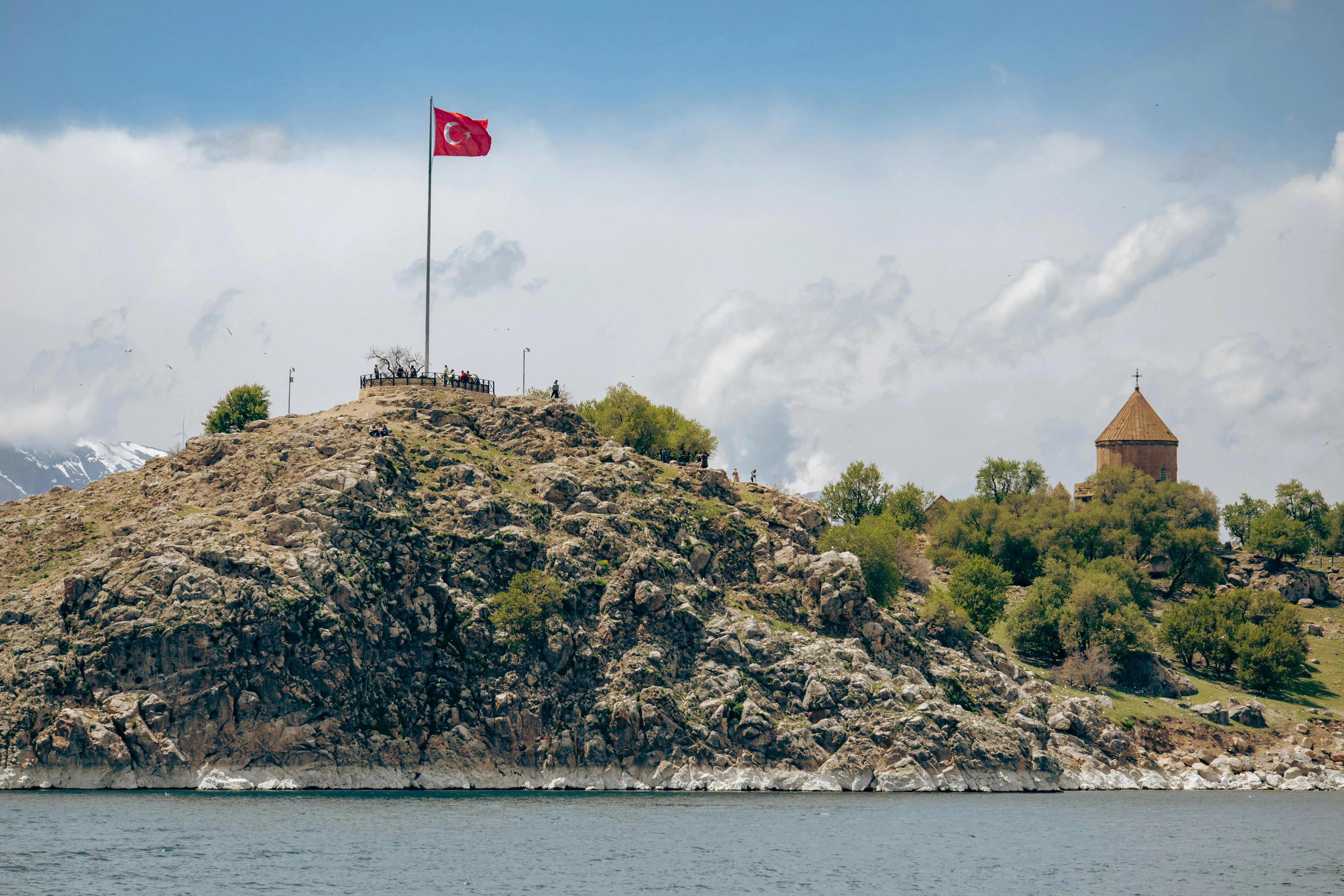 A rocky hill with a Turkish flag and a historic church, overlooking a blue lake.