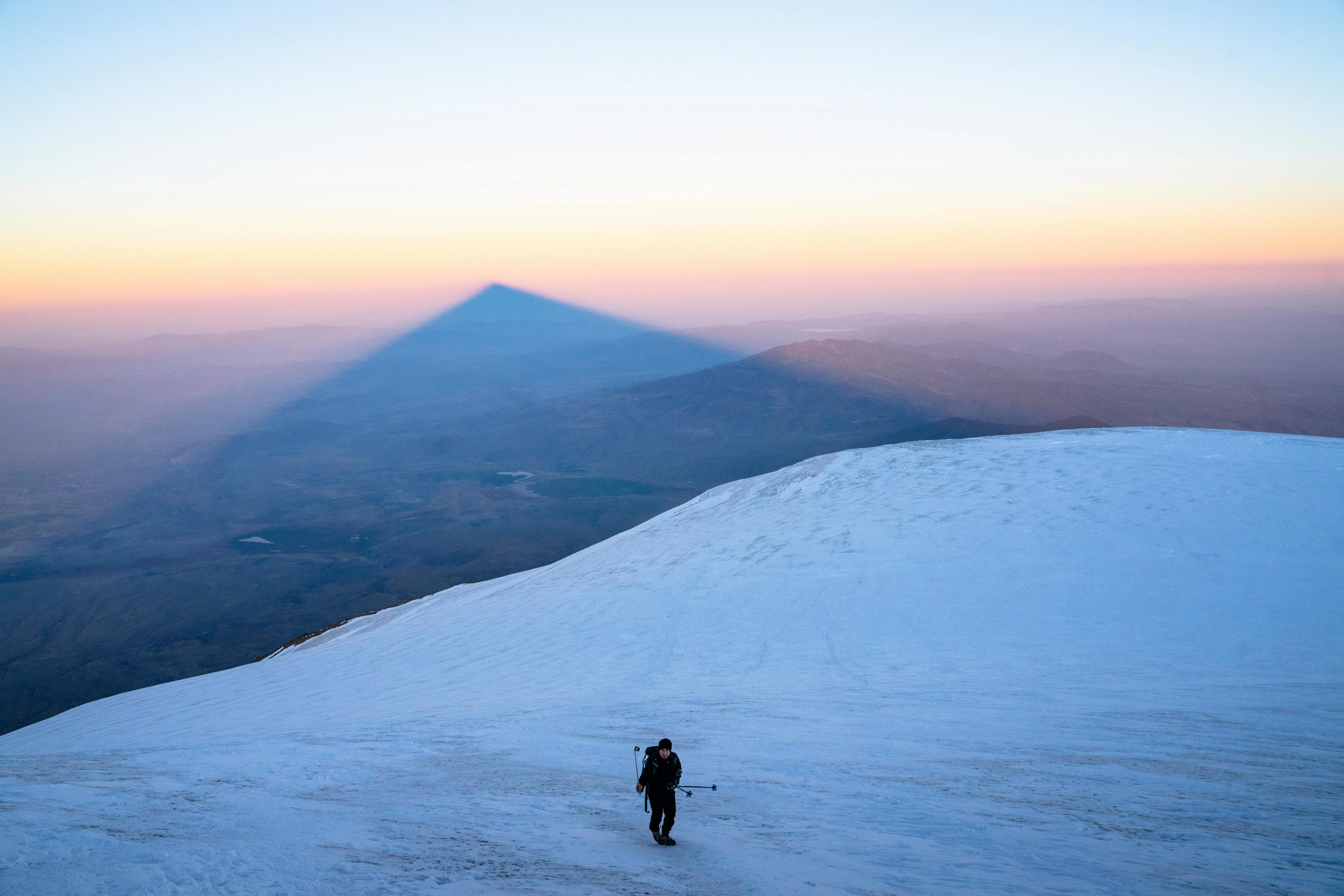 A climber walks on Mount Ararat at sunrise, with the mountain’s triangular shadow in the distance.