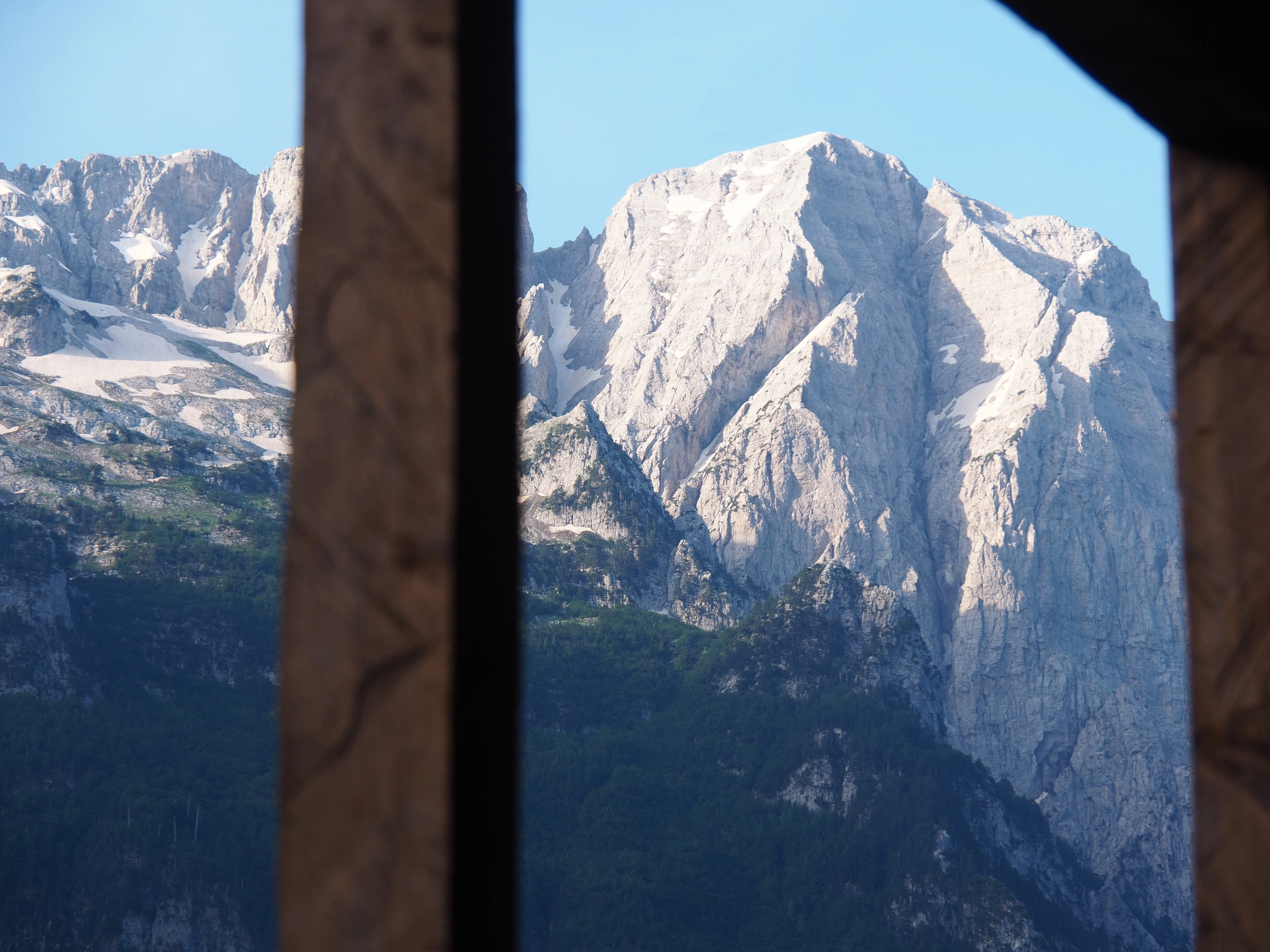 A view of snow-capped peaks in Valbona Valley, framed by the wooden beams of a traditional Albanian guesthouse.