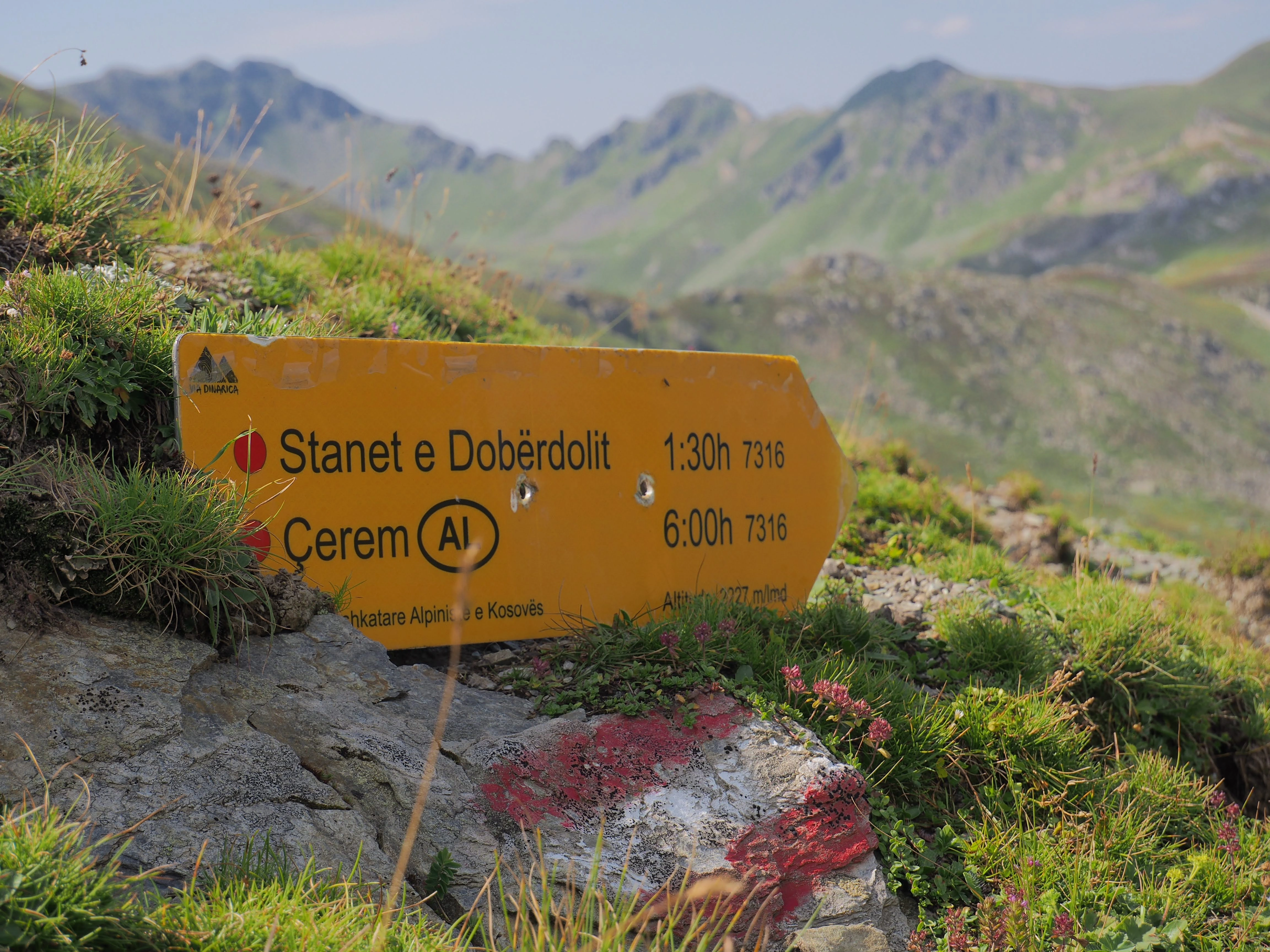 A yellow trail sign marking hiking routes in the Dobërdol Valley, set against rolling green alpine slopes.