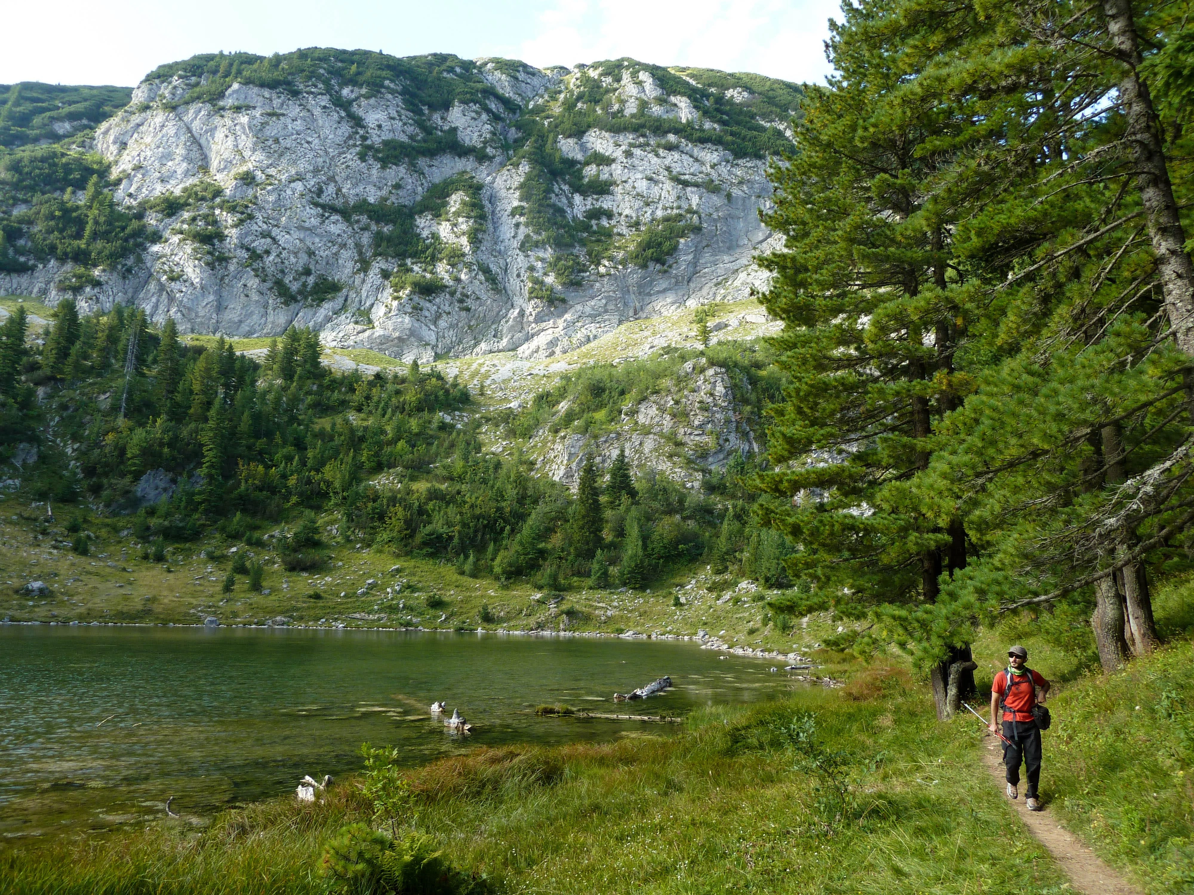 A hiker walks beside a peaceful alpine lake, surrounded by towering limestone cliffs and lush greenery.