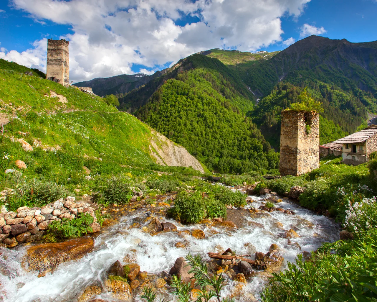 A clear rushing stream flows past ancient Svan towers and lush greenery in a remote valley in Georgia.