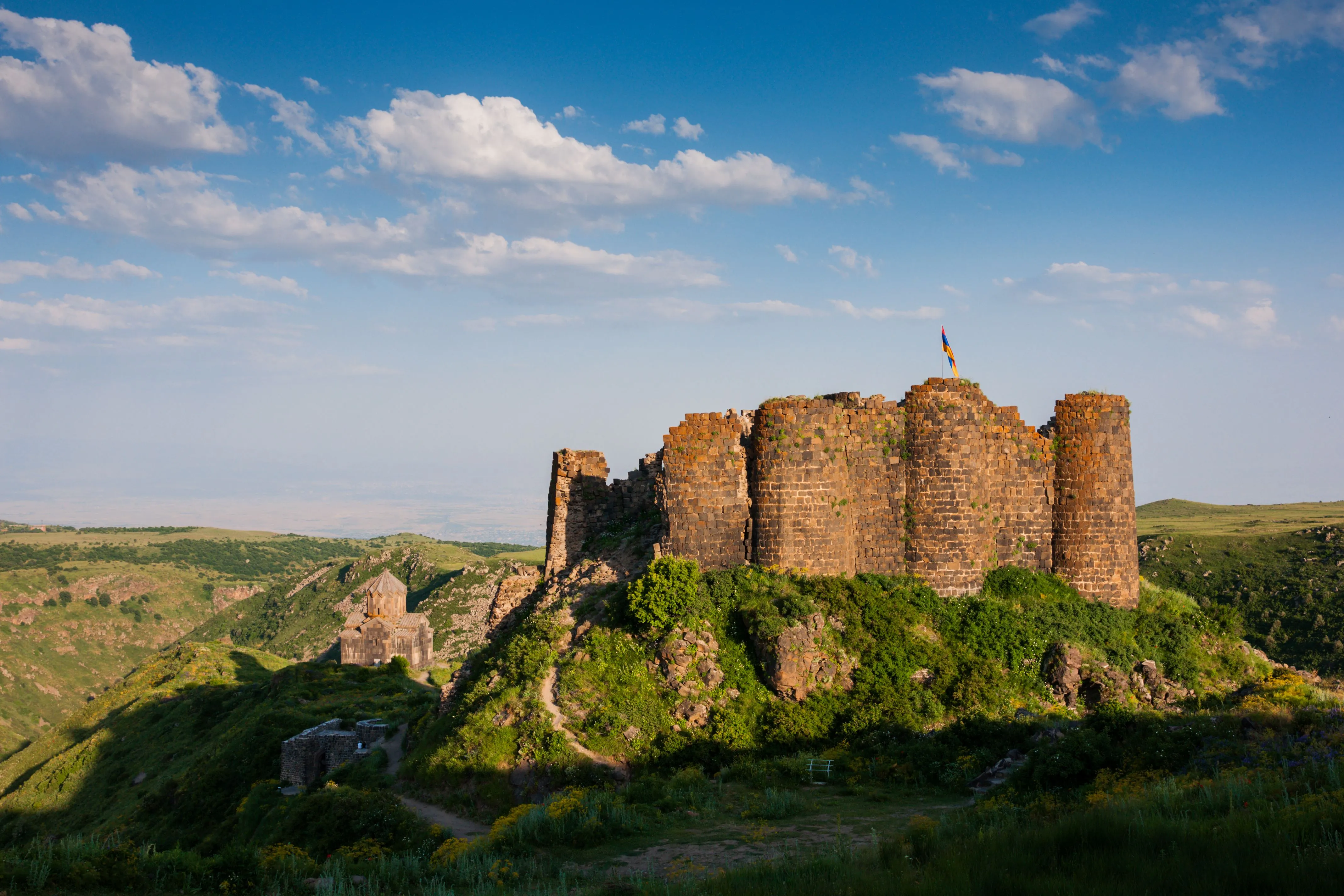 Ancient Amberd Fortress in Armenia, perched on a lush green hill under a blue sky, with an Armenian flag waving atop