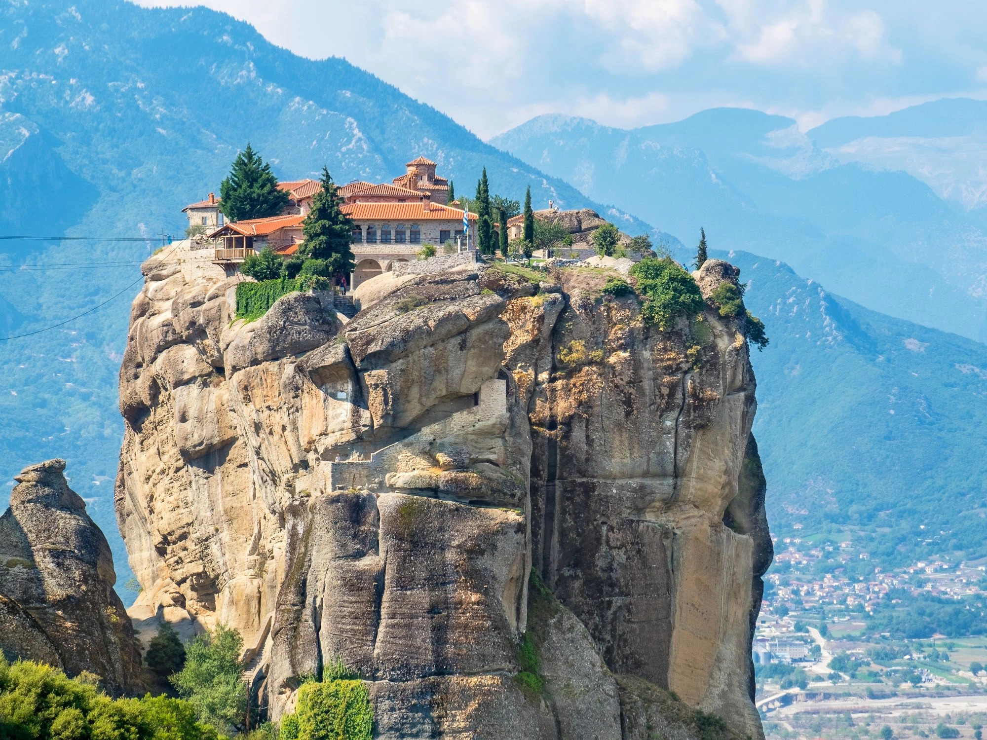 Monastery perched on massive rock cliff in Meteora Greece with blue mountains in background.