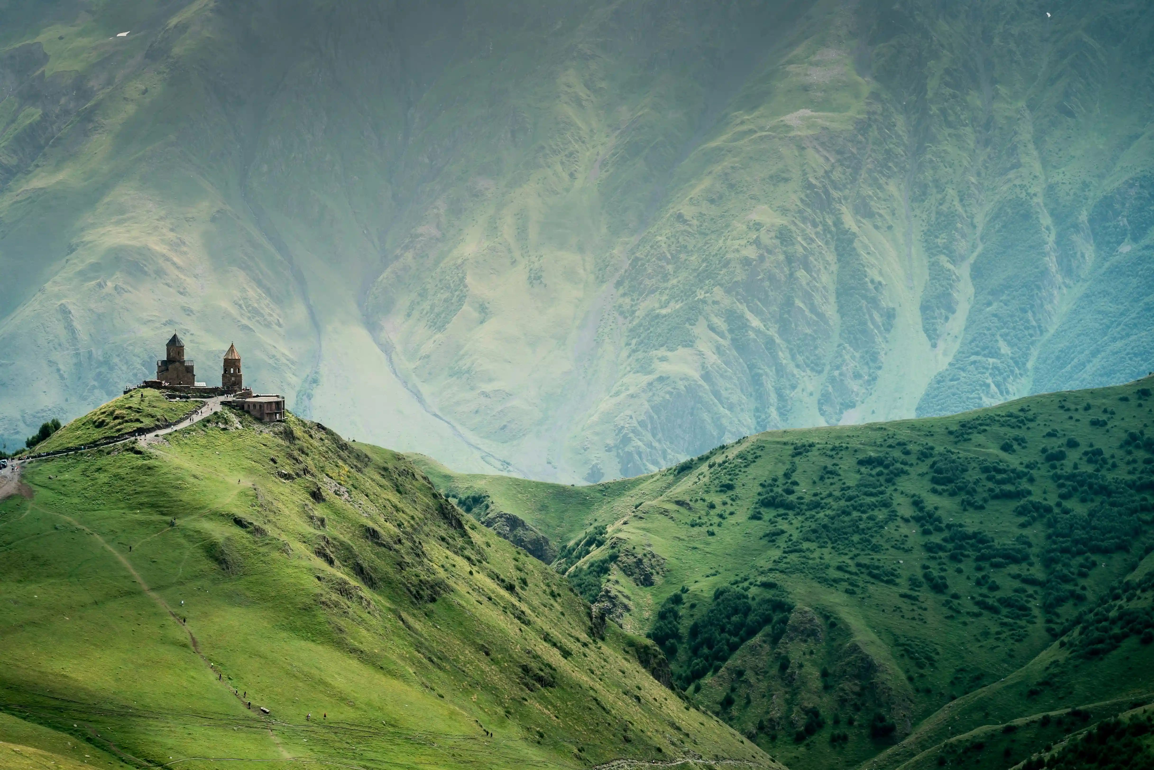 Gergeti Trinity Church atop a green hill in Georgia, surrounded by towering misty mountains.