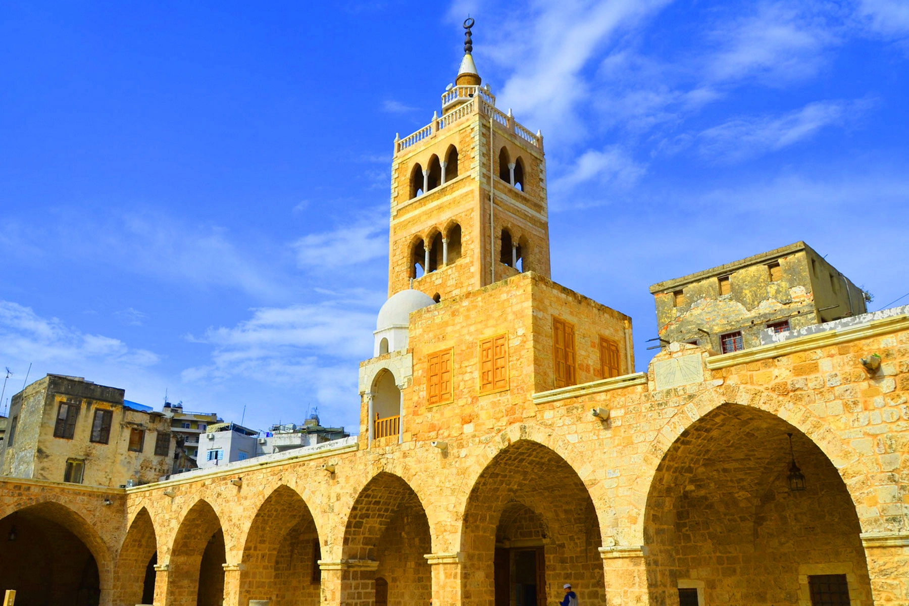 Historic mosque courtyard in Tripoli with arched walkways and a tall minaret against blue skies.