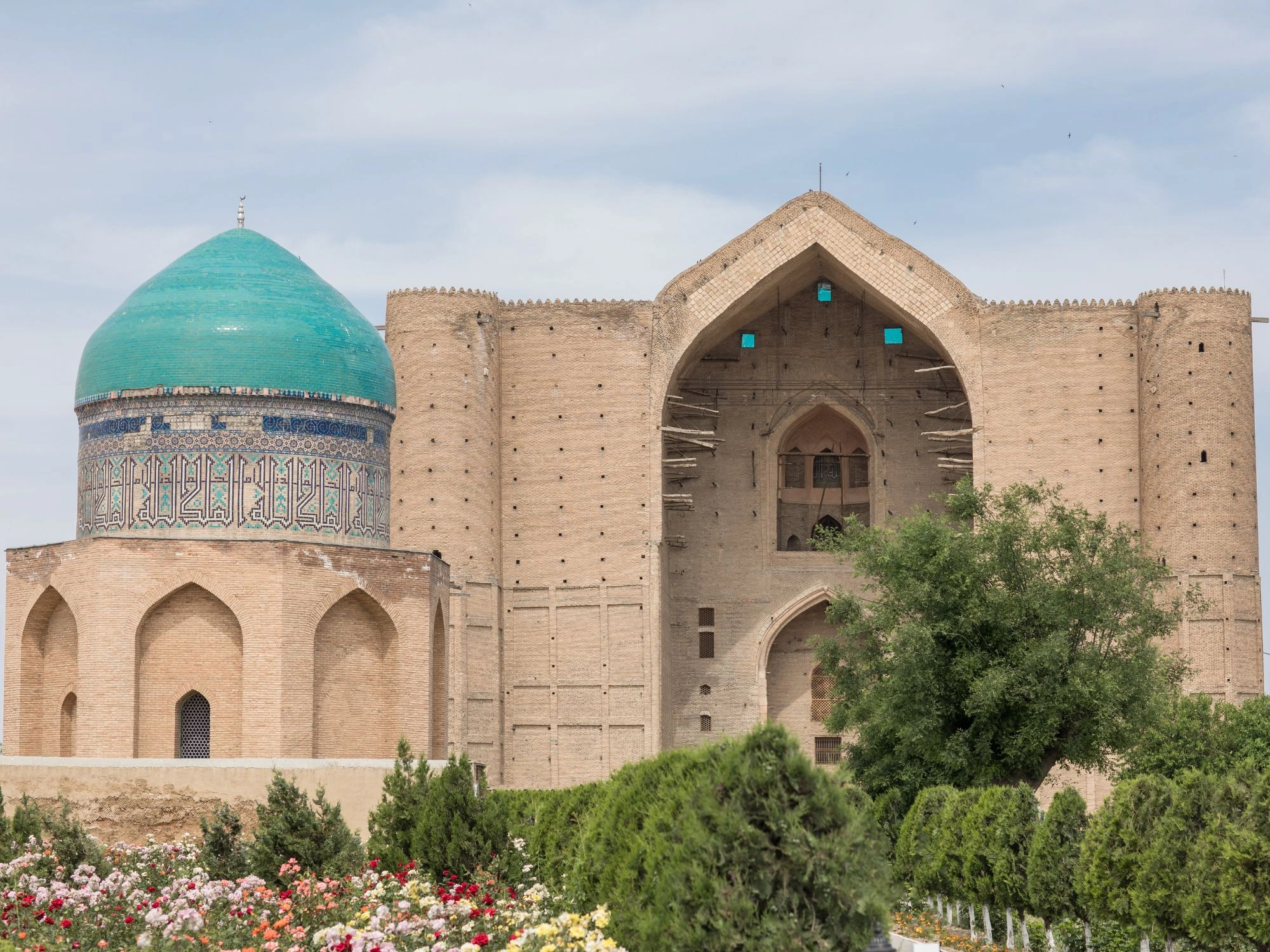 Historic mosque with turquoise dome and arched entrance in Turkistan, surrounded by gardens.