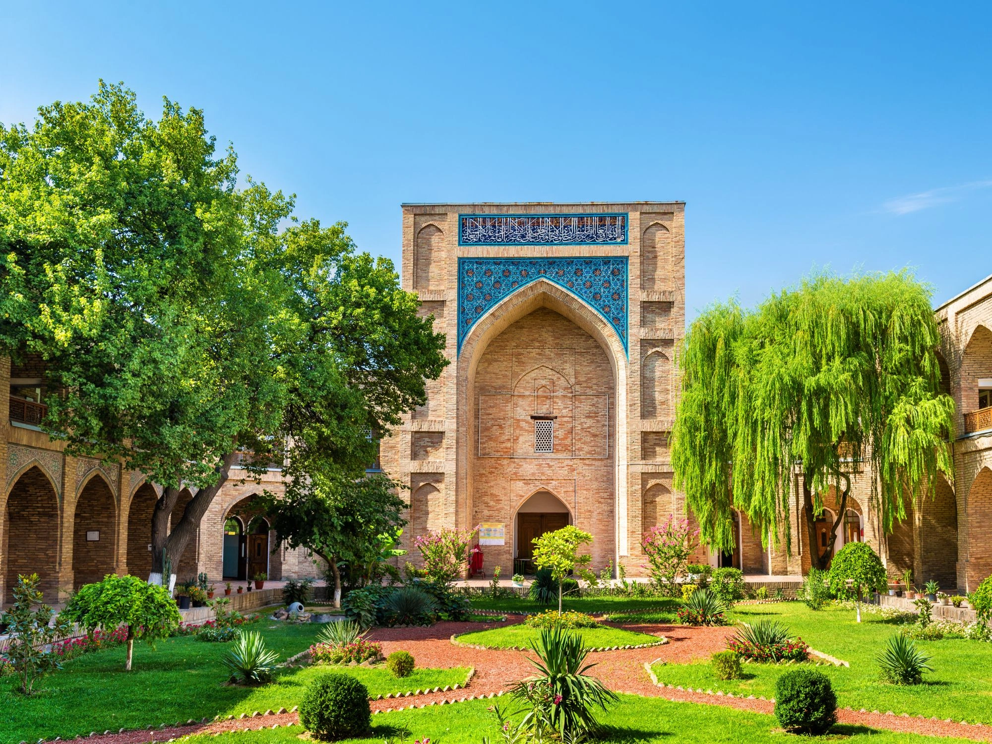 Lush garden courtyard of a historic medieval madrasa in Tashkent, Uzbekistan, showcasing Islamic architecture, arched walkways, and intricate tilework under a clear blue sky.