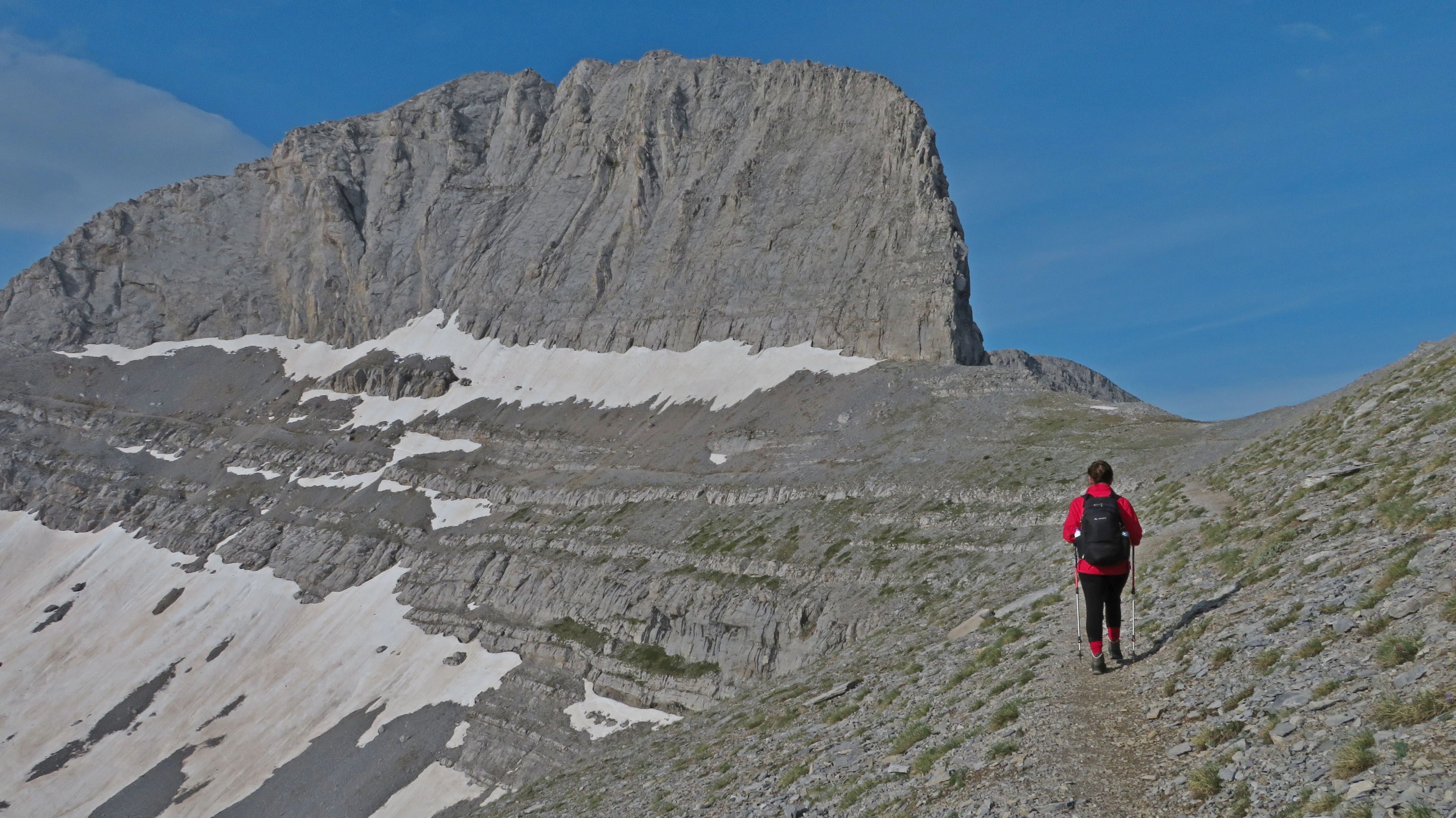 Hiker trekking below Stefani, the Throne of Zeus, along a rocky trail on Mount Olympus, Greece, with snow patches visible.