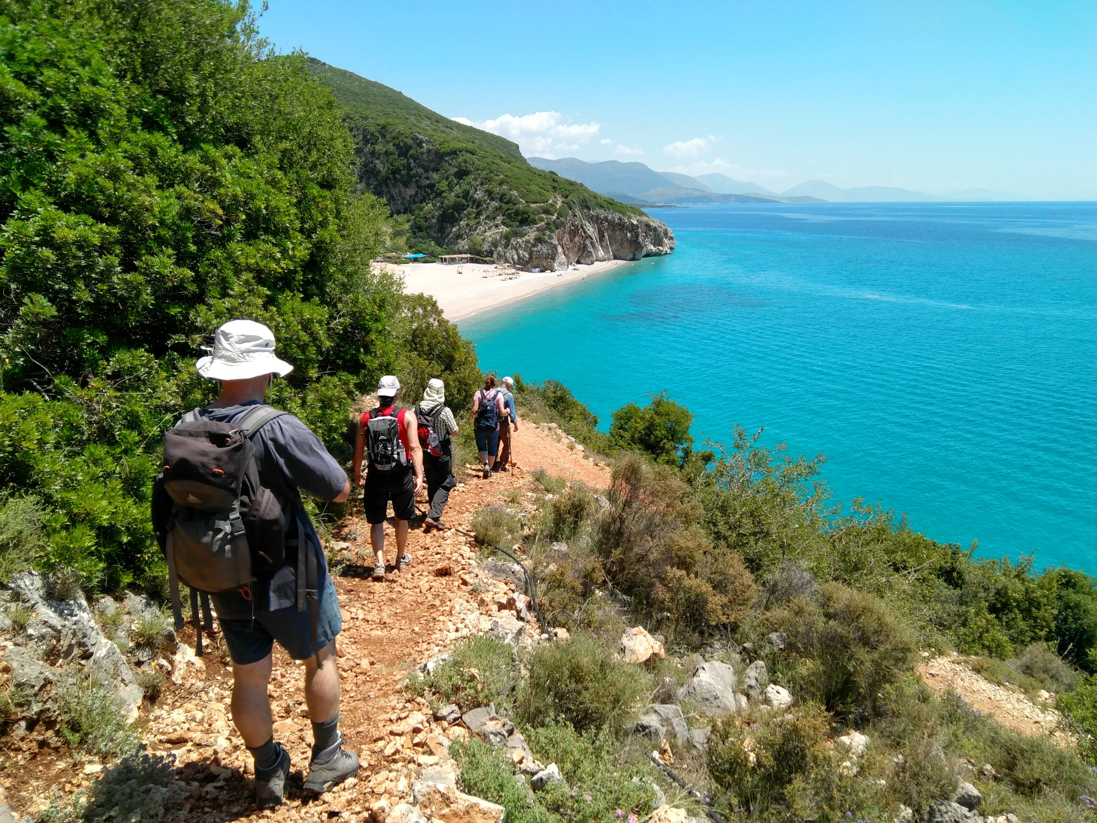 Hikers descend a rugged coastal trail toward a secluded sandy beach with crystal-clear turquoise waters in Albania.
