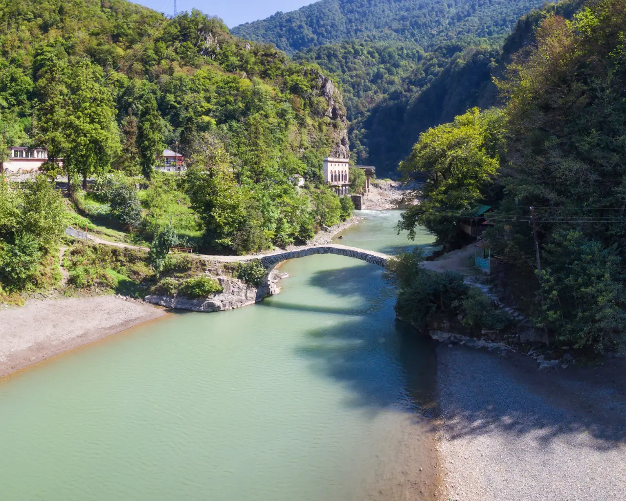 A scenic river winding through lush green mountains in Makhuntseti, Georgia, with a stone bridge crossing over the water.