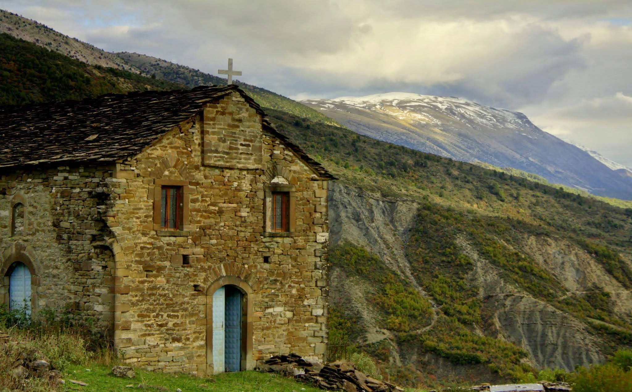 An old stone church with a wooden roof and a cross, standing on a mountain ridge with snowy peaks in the distance.