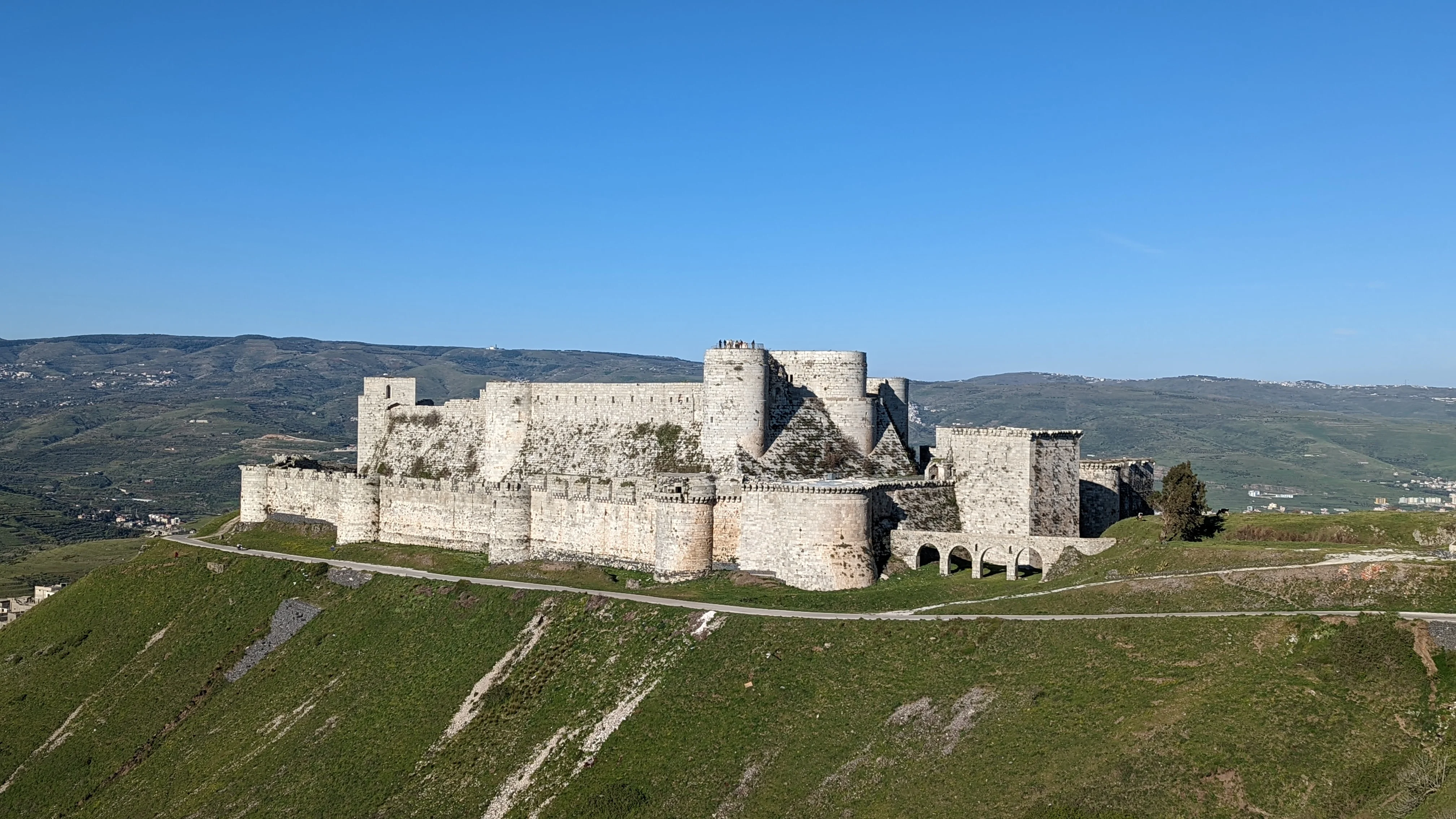 Distant view of Krak des Chevaliers fortress atop a green hill, surrounded by rolling countryside