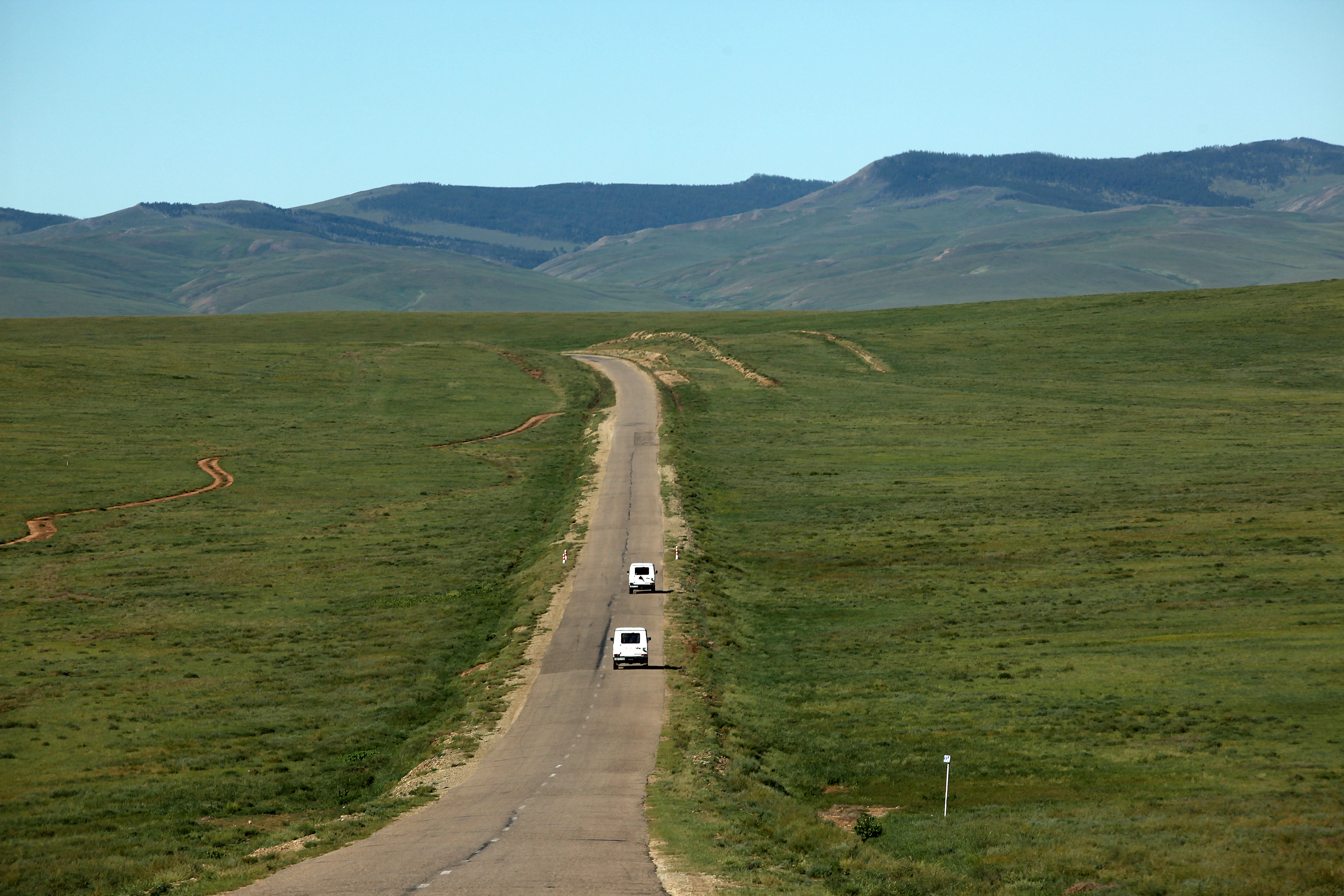 Long paved road crossing Mongolia grasslands with green hills and white vans driving ahead.
