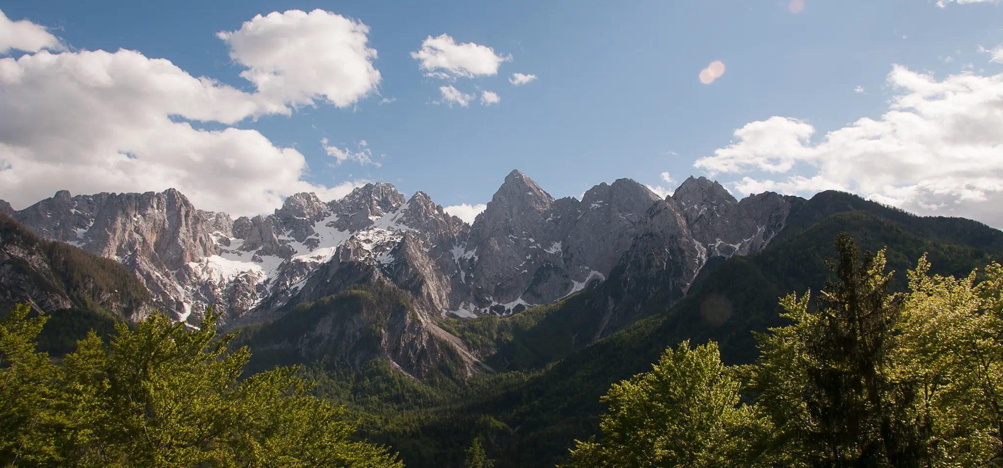 Jagged peaks of the Julian Alps with green forest below, white clouds drifting across a blue sky.