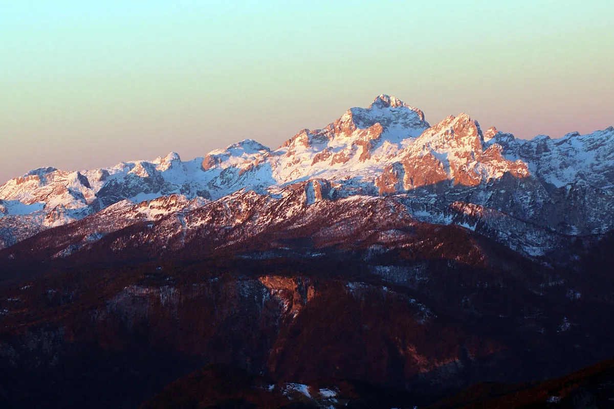 A majestic view of the Julian Alps at sunrise, with snow-covered peaks glowing in soft pink and golden hues.