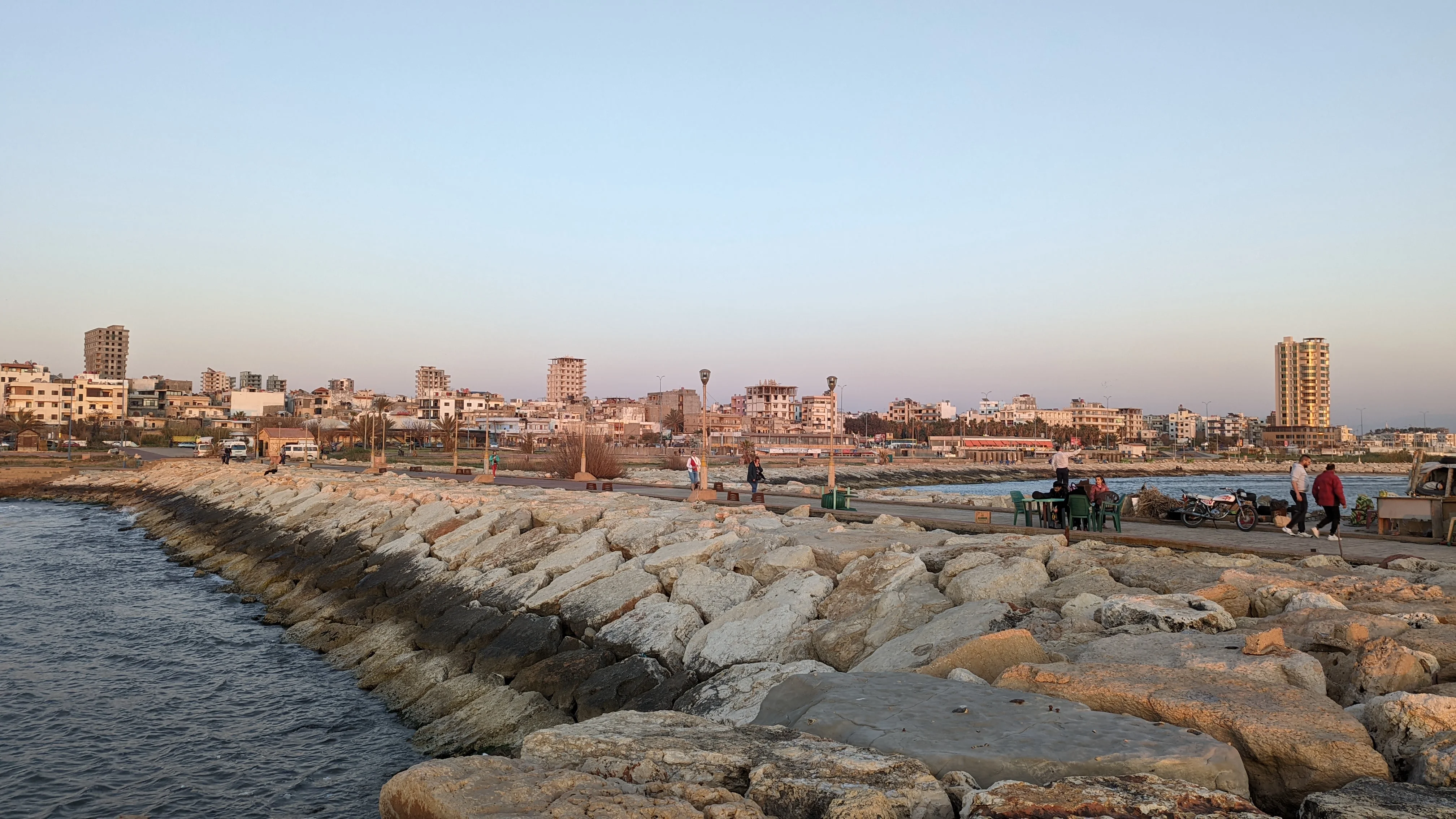 Seafront promenade in Tartus, Syria with people walking along rocky shoreline at sunset