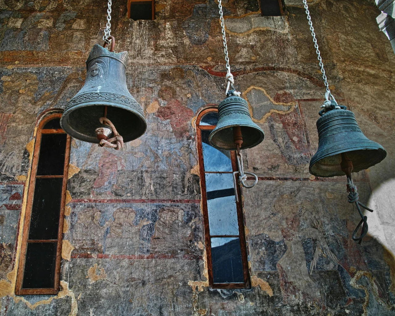 Georgian church bells – three bronze church bells hanging by chains against a frescoed wall and arched window.