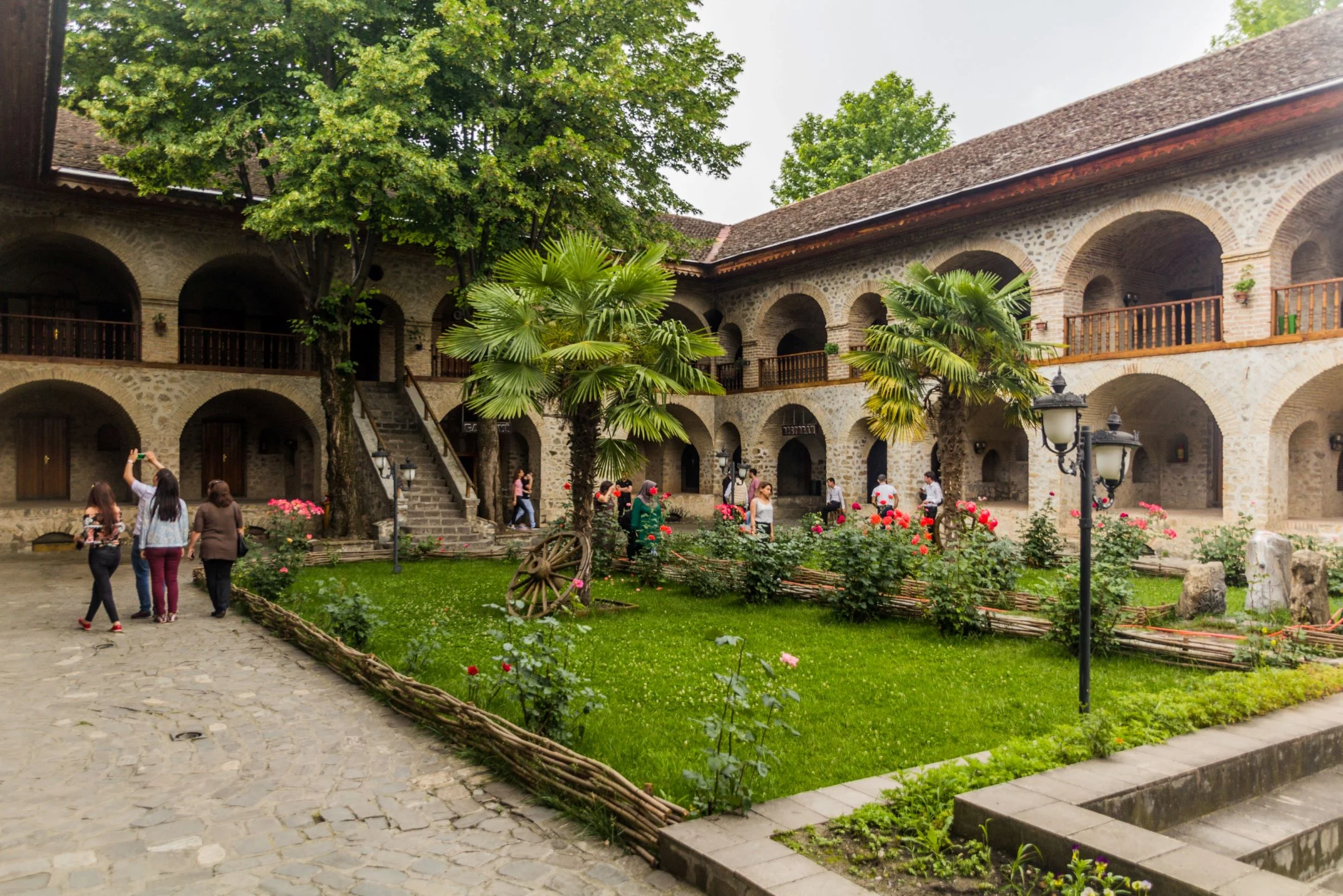 Sheki caravanserai courtyard – inner courtyard of the Sheki caravanserai with two‑story stone arcades, lush garden and visitors exploring.