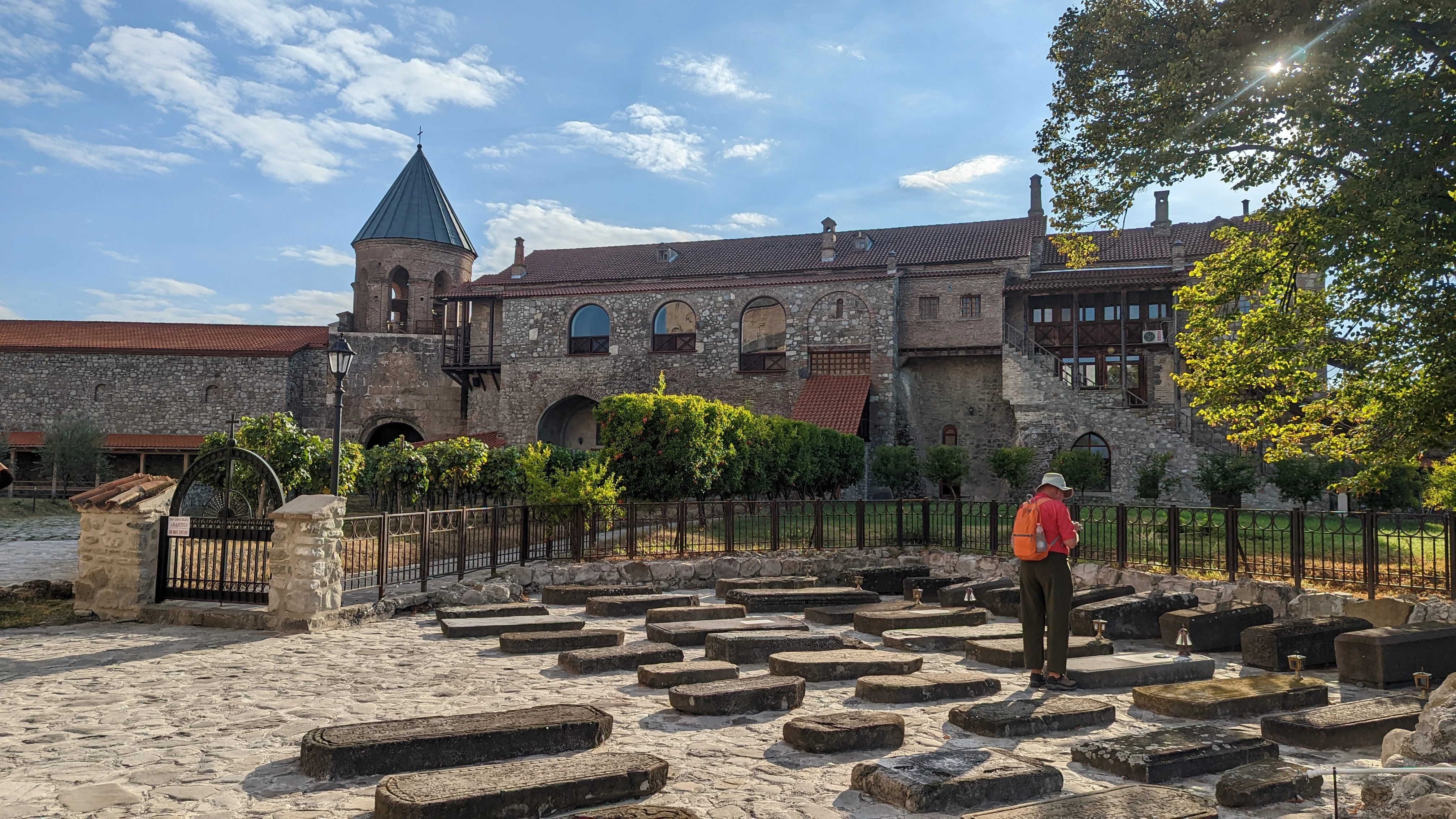 Georgian monastery cemetery – traveler walking among old stone graves at a historic monastery complex with bell tower and red tiled roofs.