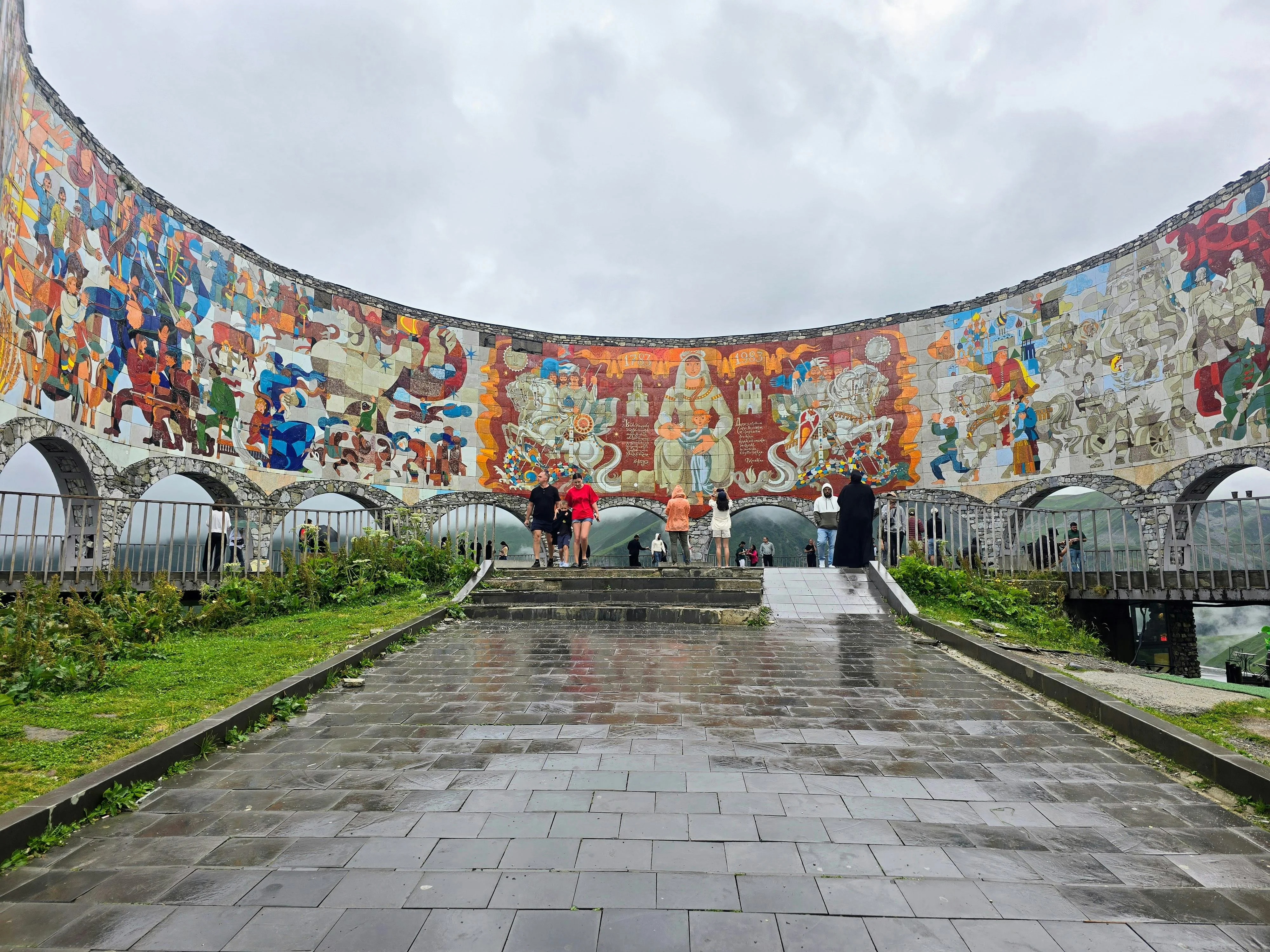 Friendship Monument Georgia – panoramic view from the Soviet‑era Friendship Monument with colorful murals overlooking the Caucasus Mountains.