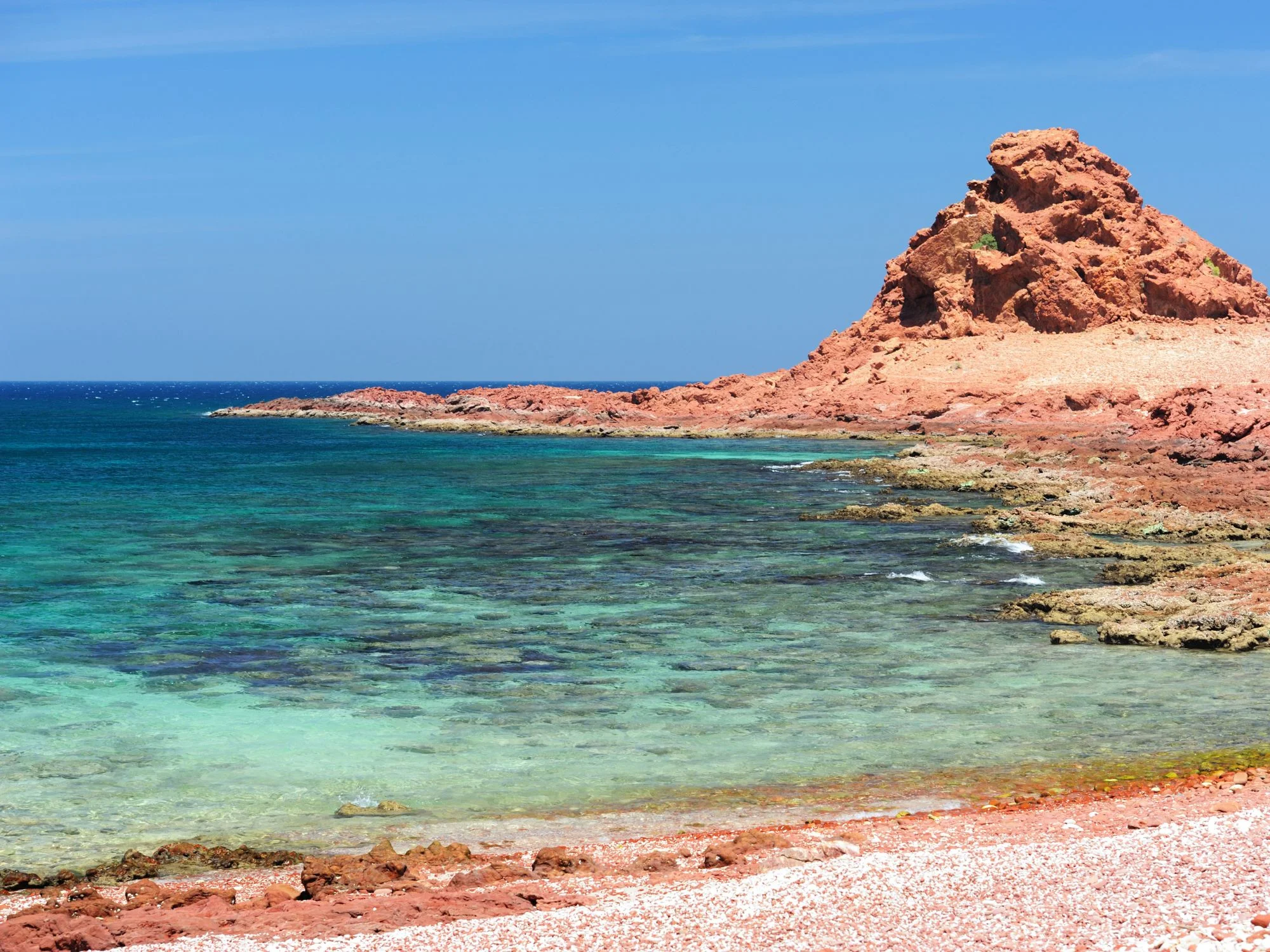 Red rocky promontory and coral beach on Socotra with clear turquoise shallows and deep blue sea.