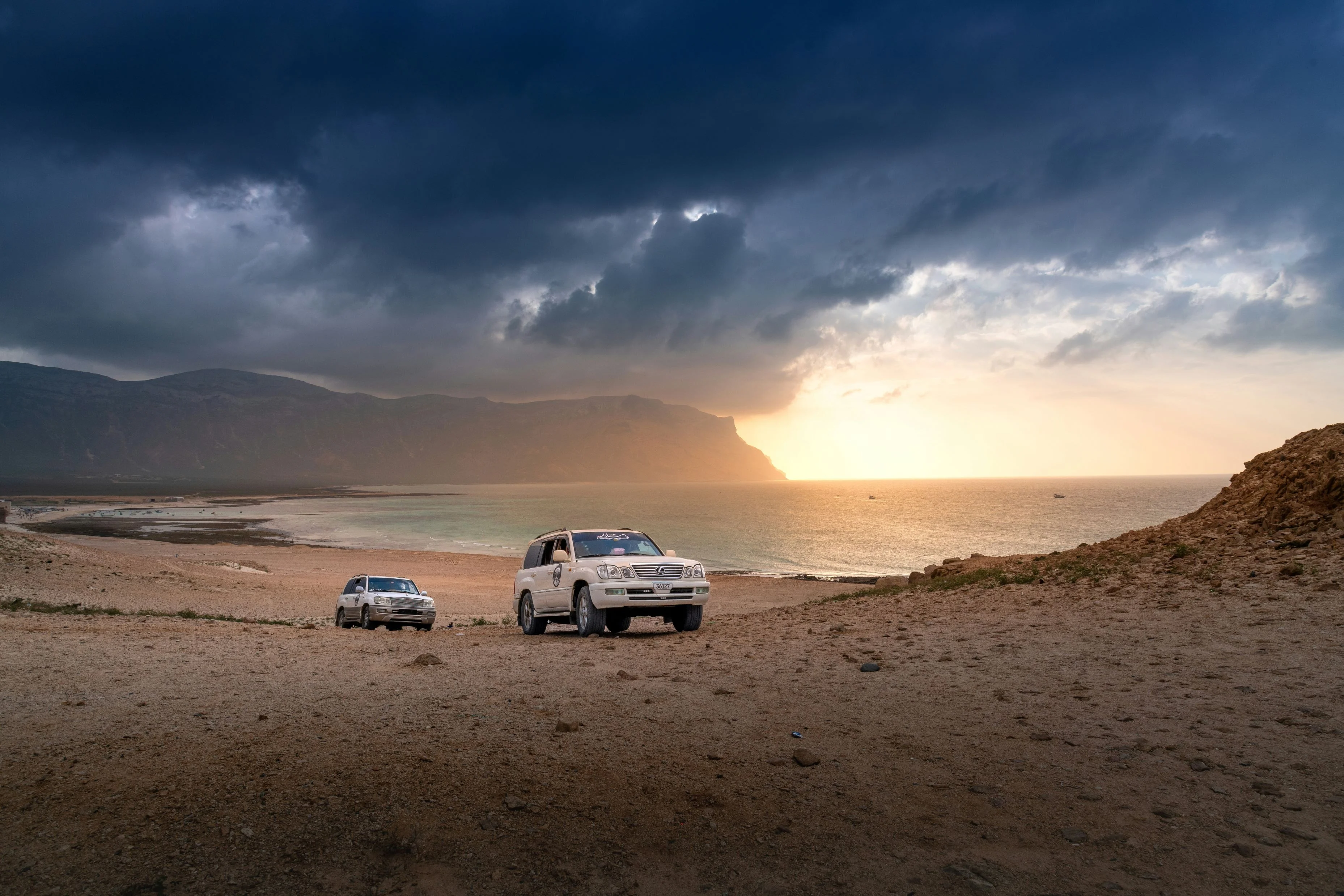 4x4 vehicles driving along sandy Socotra beach with mountains and dramatic sunset sky.